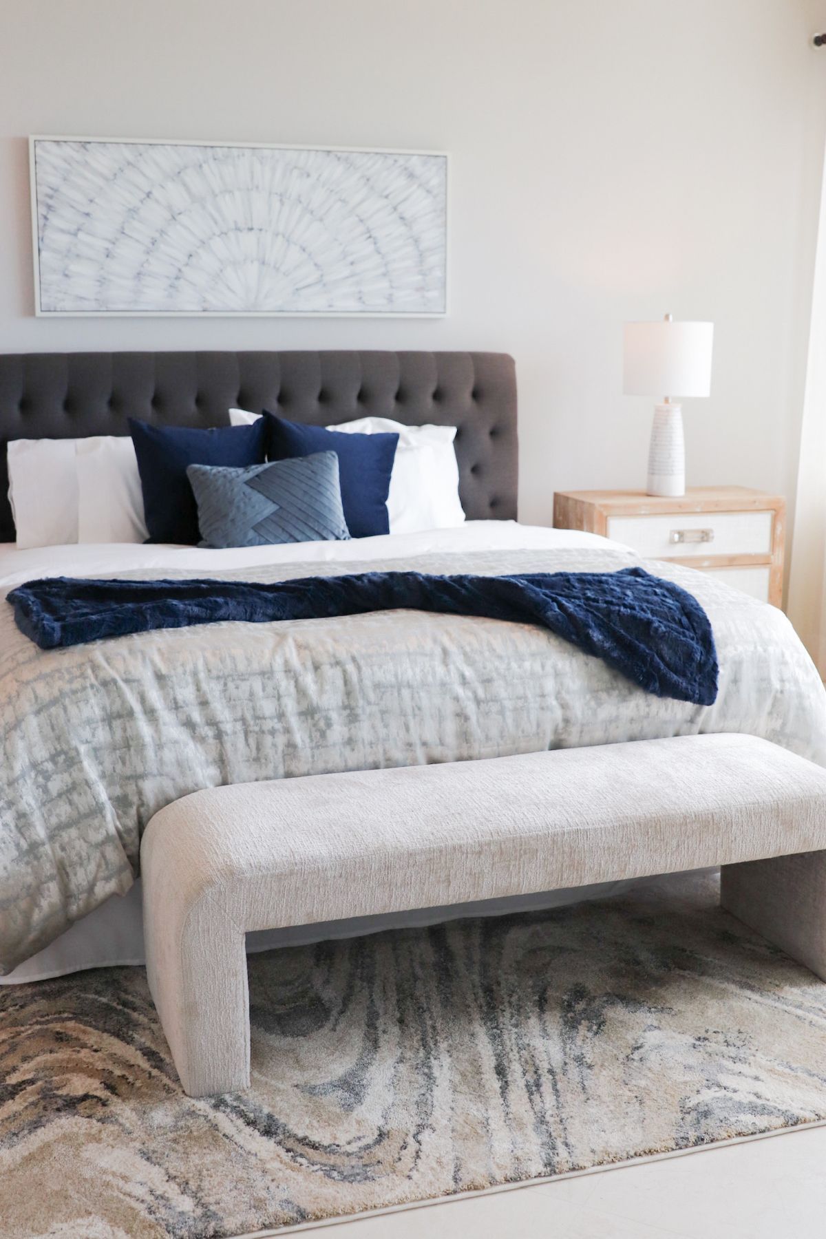 Bedroom with grey tufted headboard, blue accents, light-colored bedding, bench, and patterned rug.