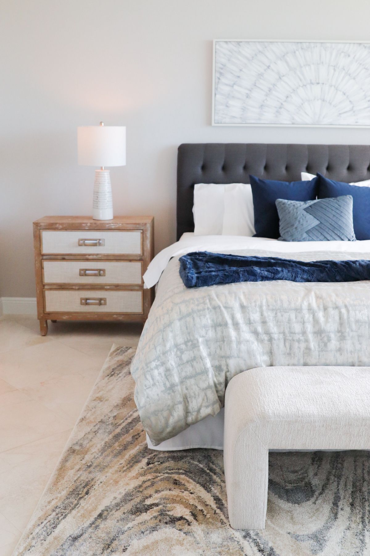 Bedroom with a tufted gray headboard, white bedding, and navy blue pillows; a wooden nightstand with a lamp.