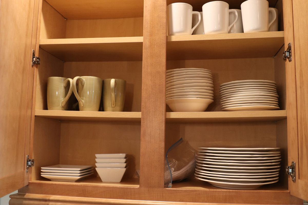 Wooden kitchen cabinet open, displaying dishes and mugs on shelves.