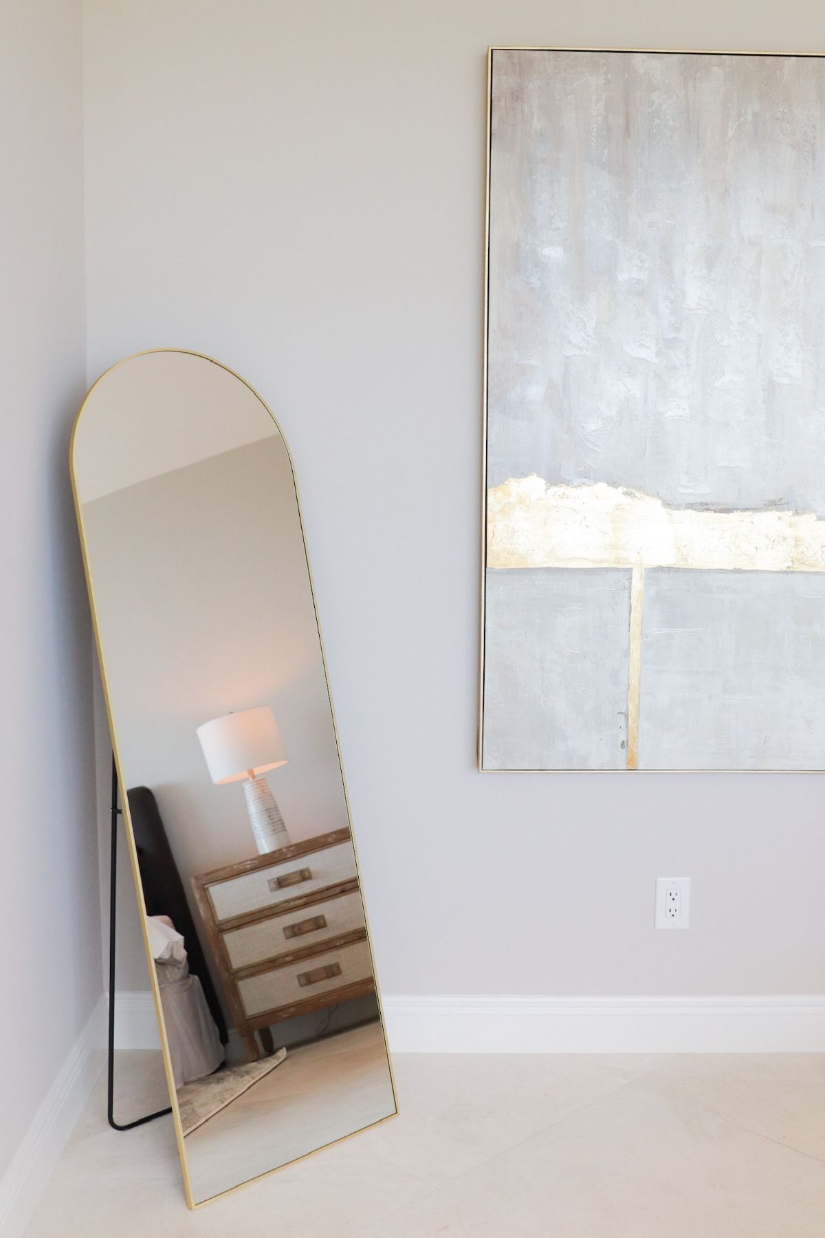 Full-length arched gold mirror propped in a corner, reflecting a bedroom with a dresser and art on the wall.