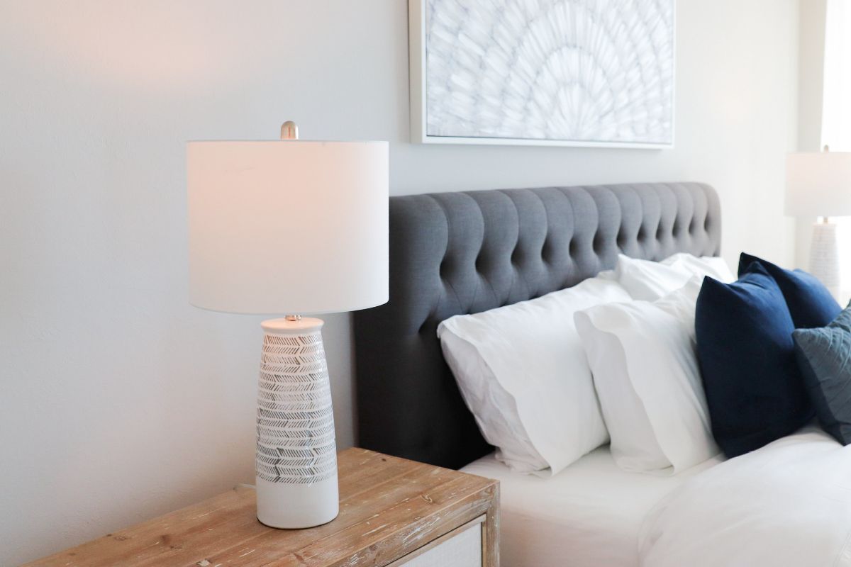 Bedroom with gray tufted headboard, white bedding, blue pillows, and bedside lamp on a wooden table.