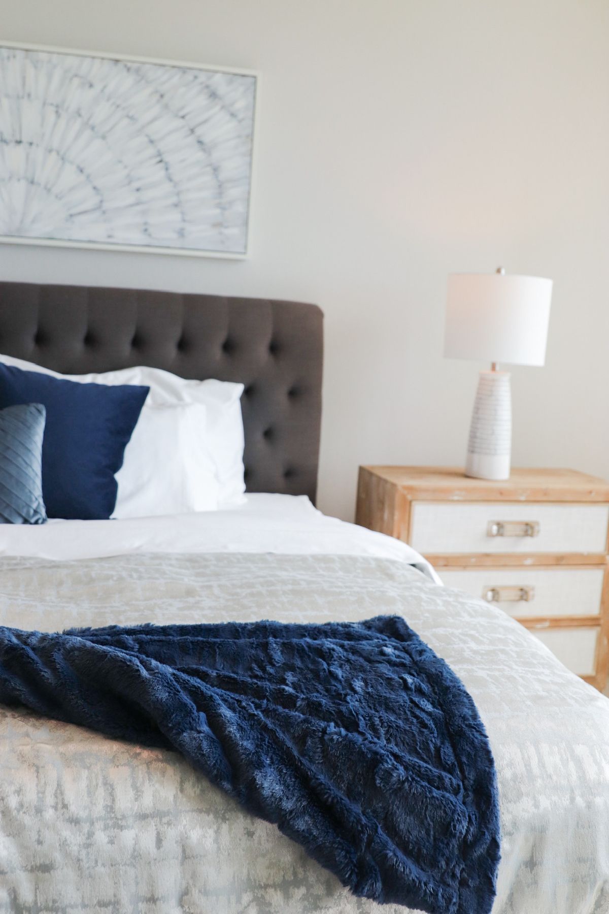 Bedroom with gray tufted headboard, white bedding, navy throw, and light wood nightstand with lamp.