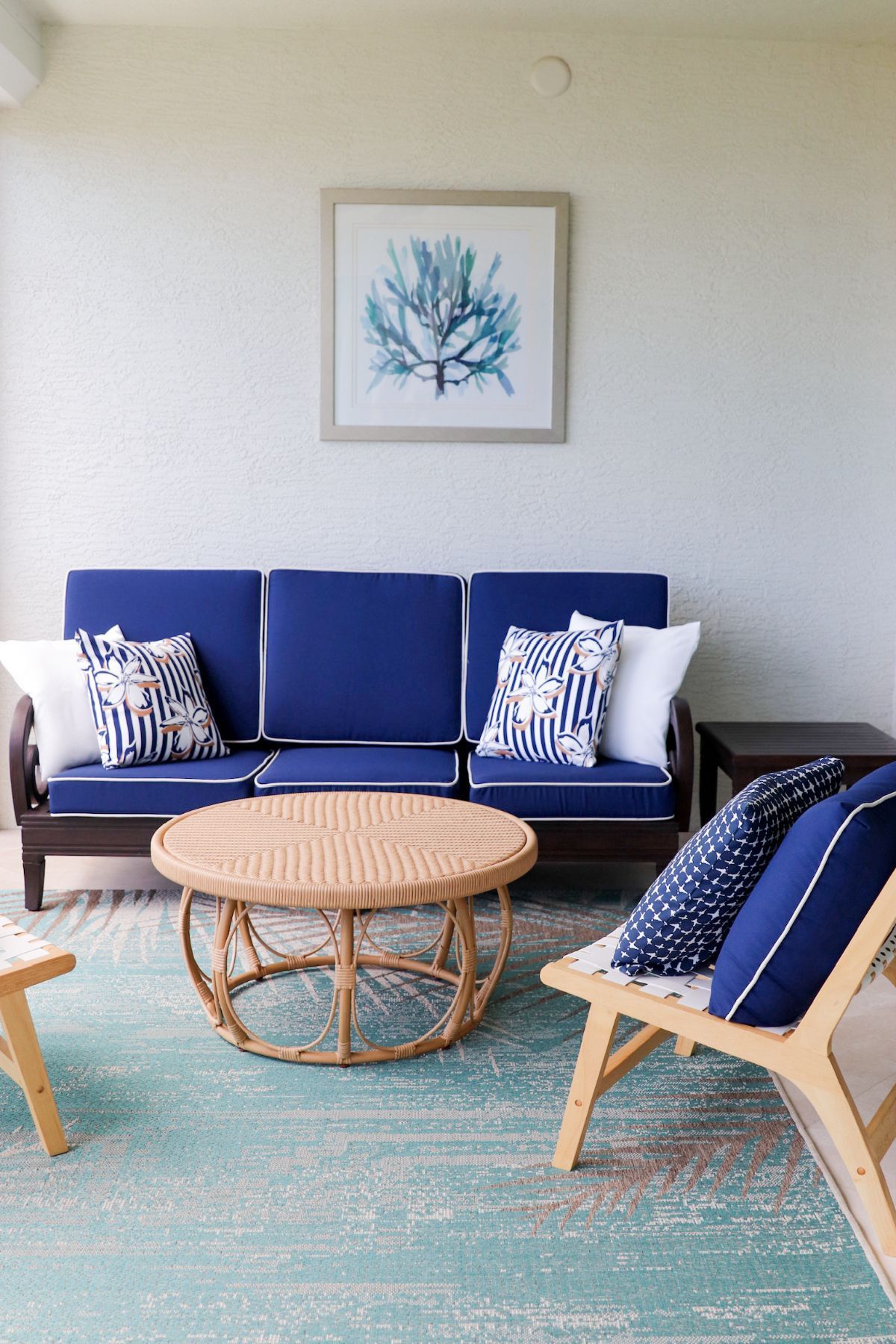 Outdoor seating area: navy blue couch with white trim, wicker coffee table, teal rug, ocean-themed artwork.