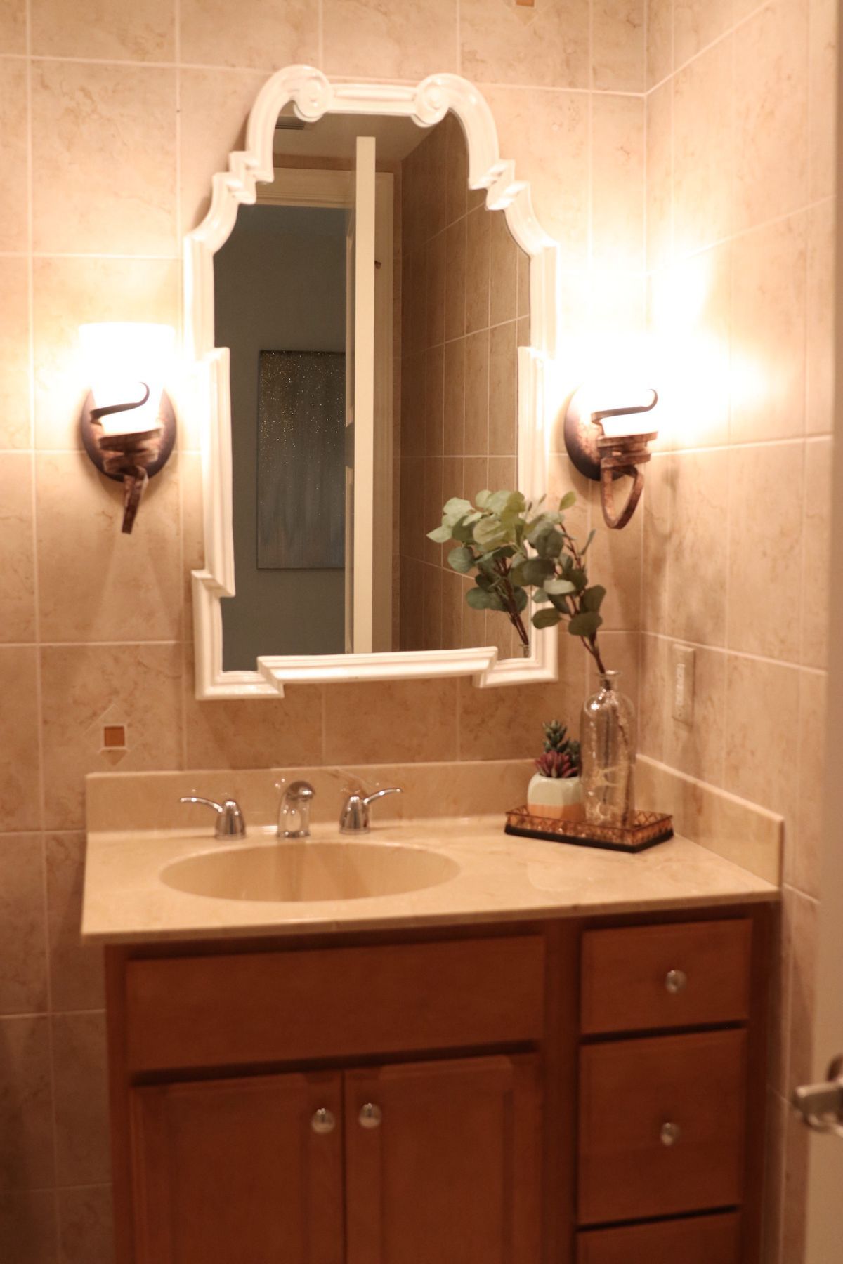 Bathroom with tan tiled walls, light wooden vanity, oval sink, and ornate white mirror.