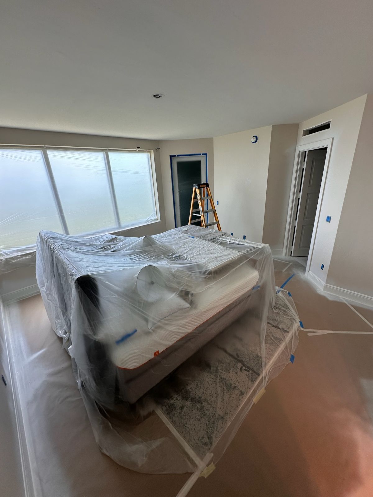 Bedroom interior during renovation; bed covered with plastic, ladder near door, new paint, and exposed floors.