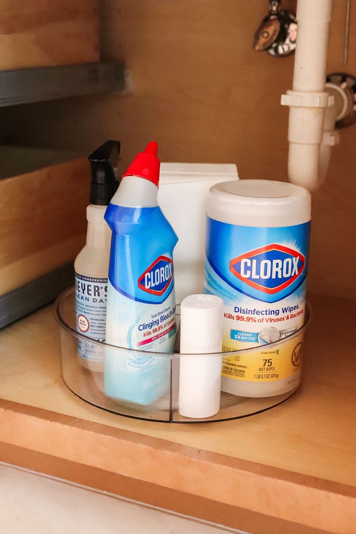 Cleaning supplies organized on a clear, rotating tray under a sink. Includes spray bottles, Clorox wipes, and cleaner.