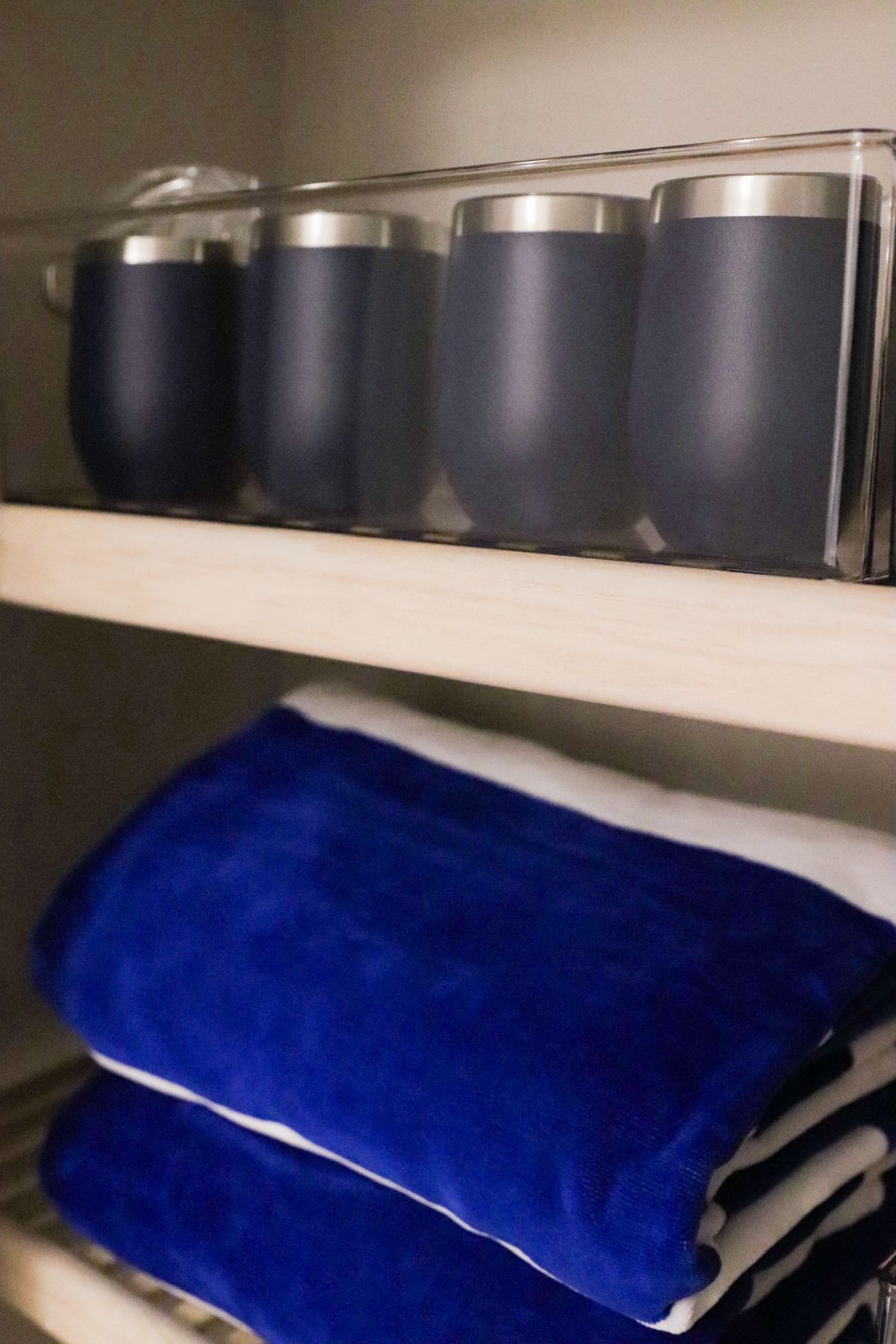 Blue insulated tumblers in a clear container on a shelf above folded blue towels.
