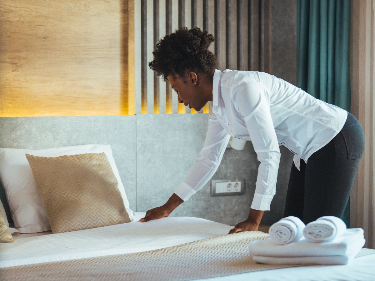 Person in a white shirt and black pants making a bed in a hotel room.