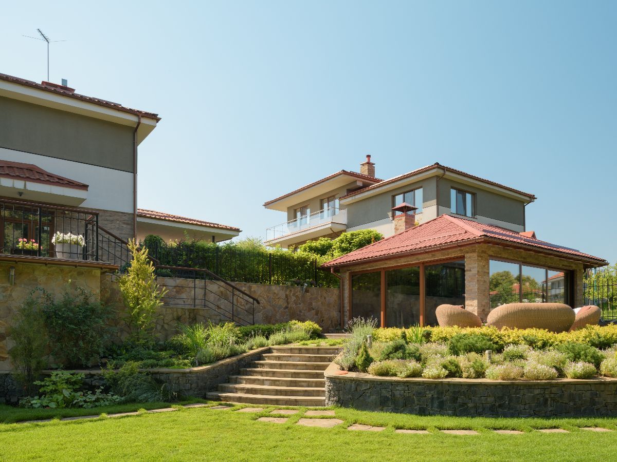 Stone steps lead up to a gazebo with a red tile roof and house. Green lawn and shrubs.