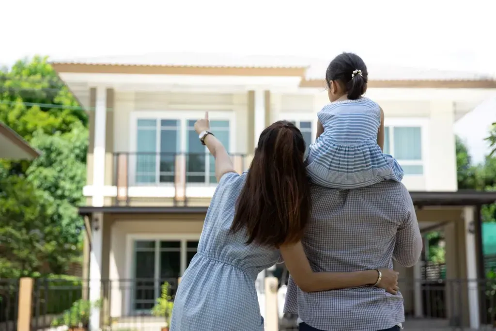 Family standing with a child on shoulders, pointing toward a two-story residential house on a sunny day.