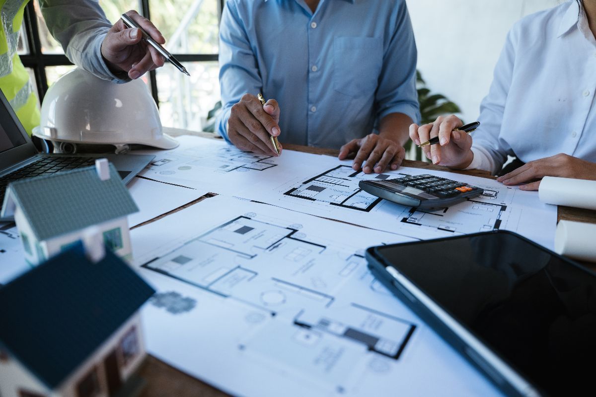 Architects and clients review blueprints and models on a wooden table, using a calculator and tablet for planning.