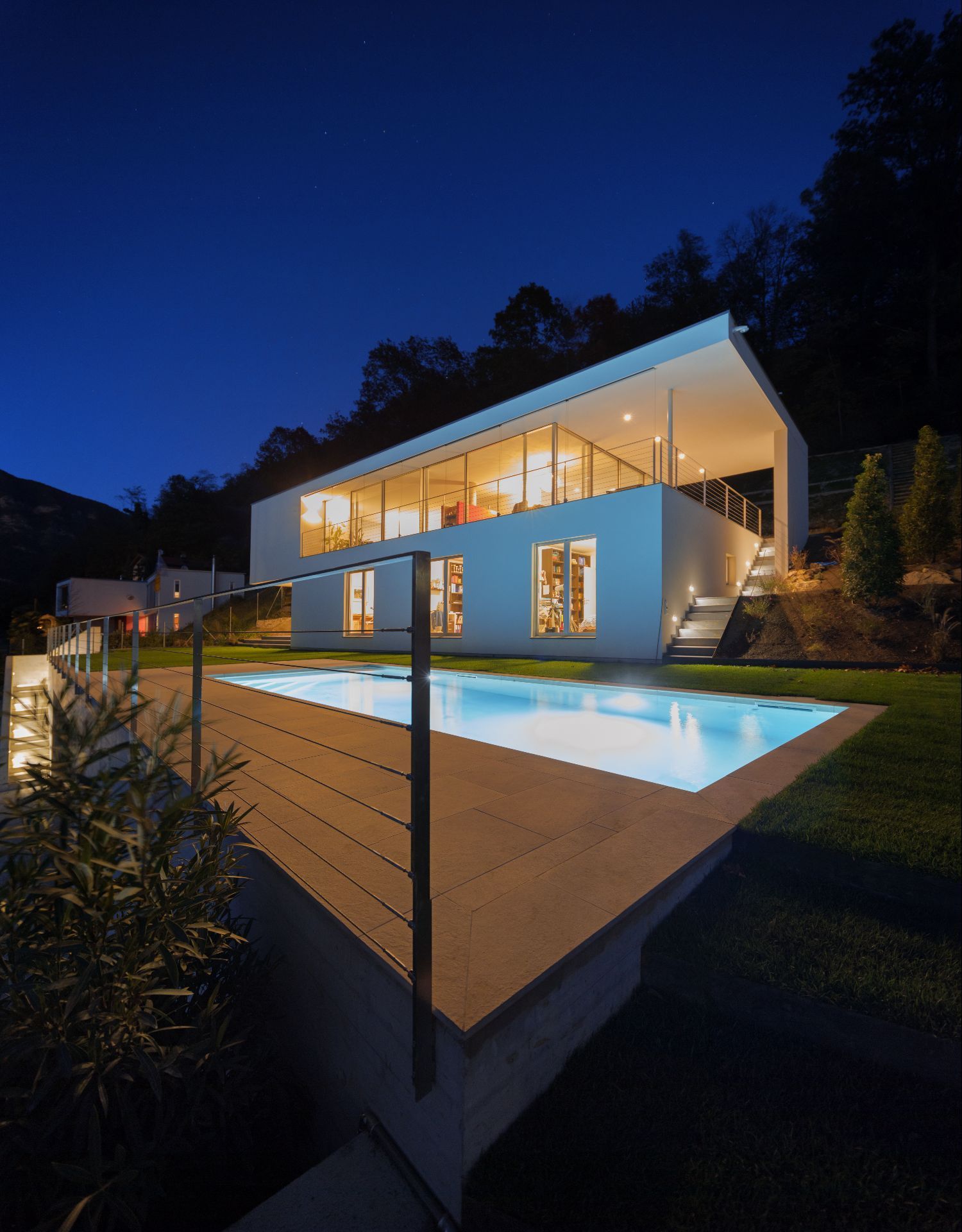 A modern white two-story house with a lit swimming pool at twilight, surrounded by a lawn and hillside trees.