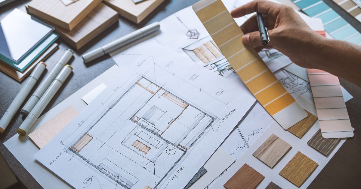 Hand holding paint swatches over interior design sketches, with art supplies on a desk.