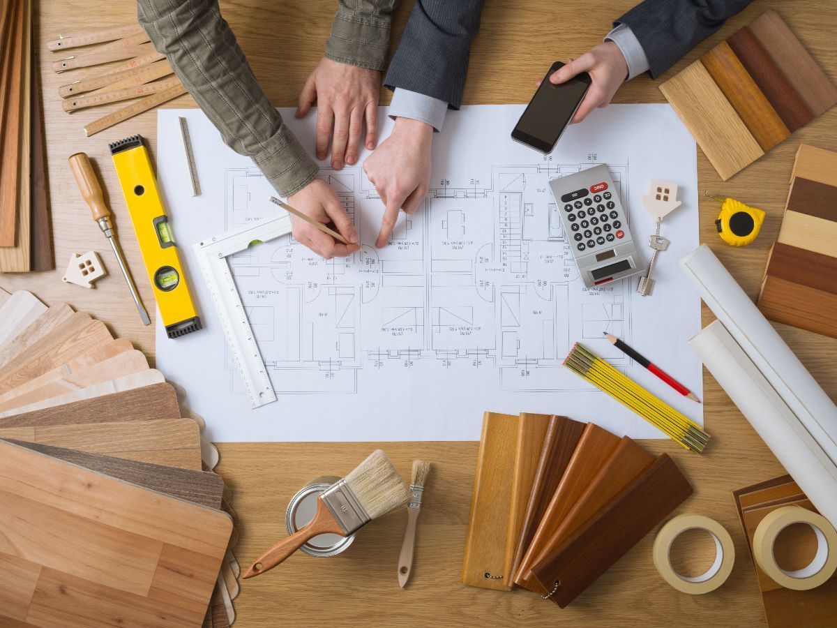 Two people reviewing floor plans on a wooden desk surrounded by flooring samples, tools, and construction supplies.