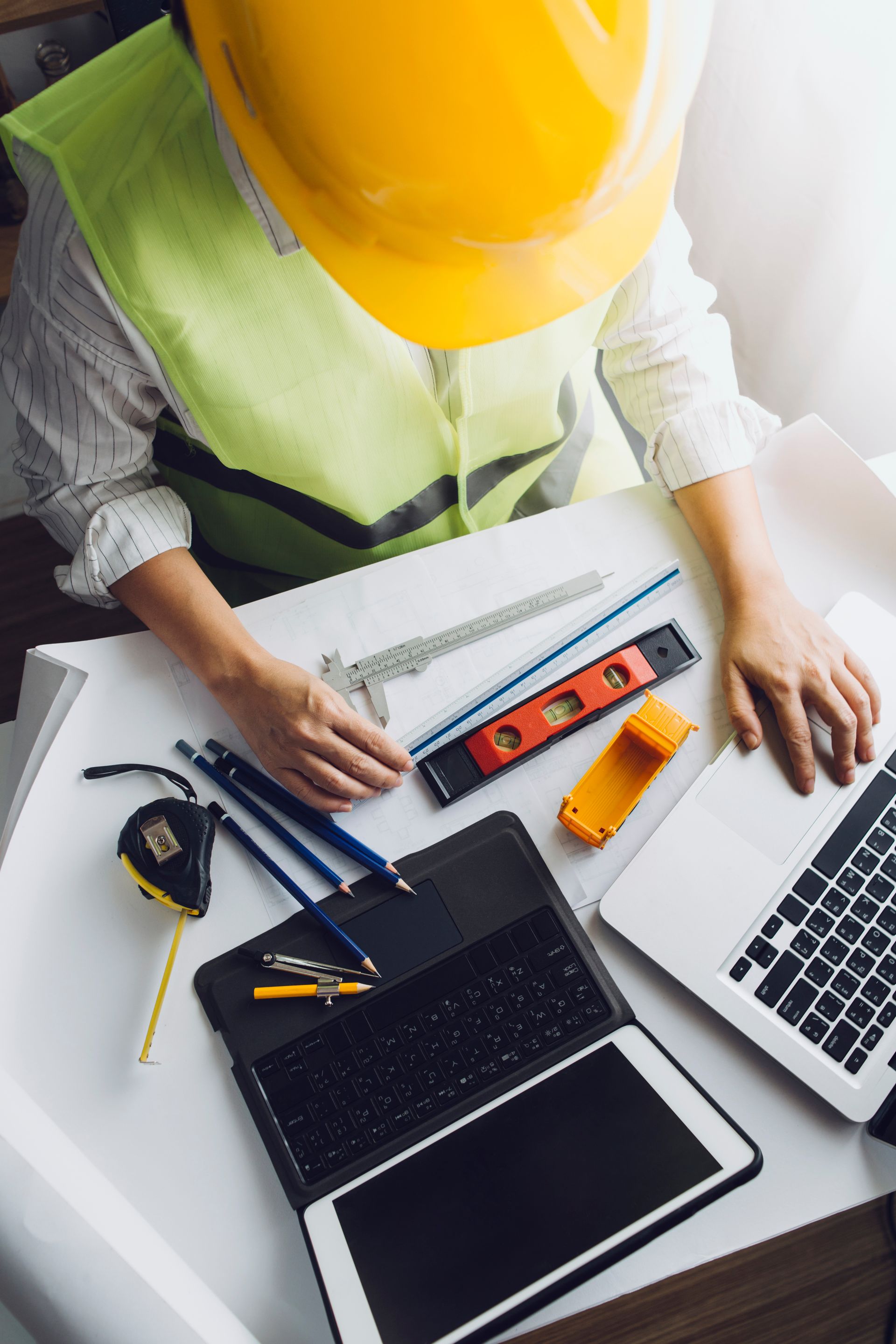 An engineer in a hard hat and safety vest works at a desk with a laptop, tablet, and various measuring tools.