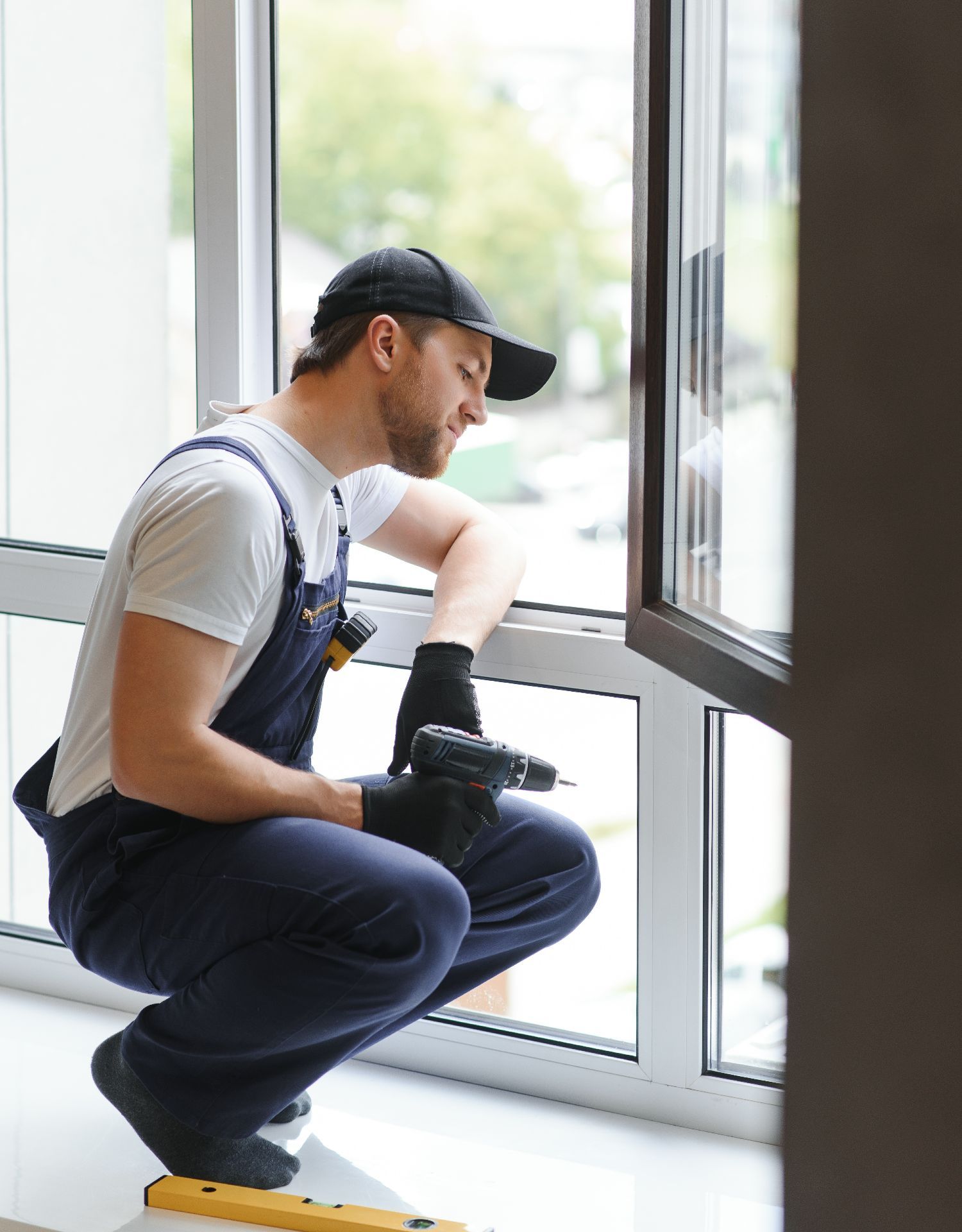 A person in workwear kneels by a window, holding a power drill while examining the window frame.