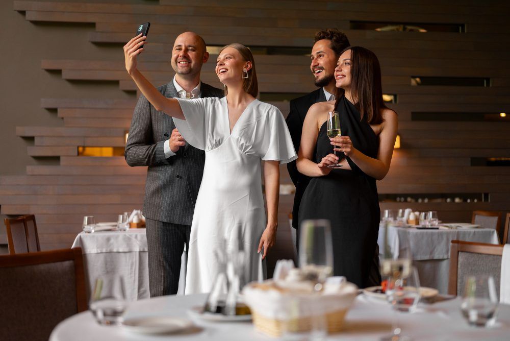 Four people taking a selfie in a restaurant. Two hold champagne glasses.