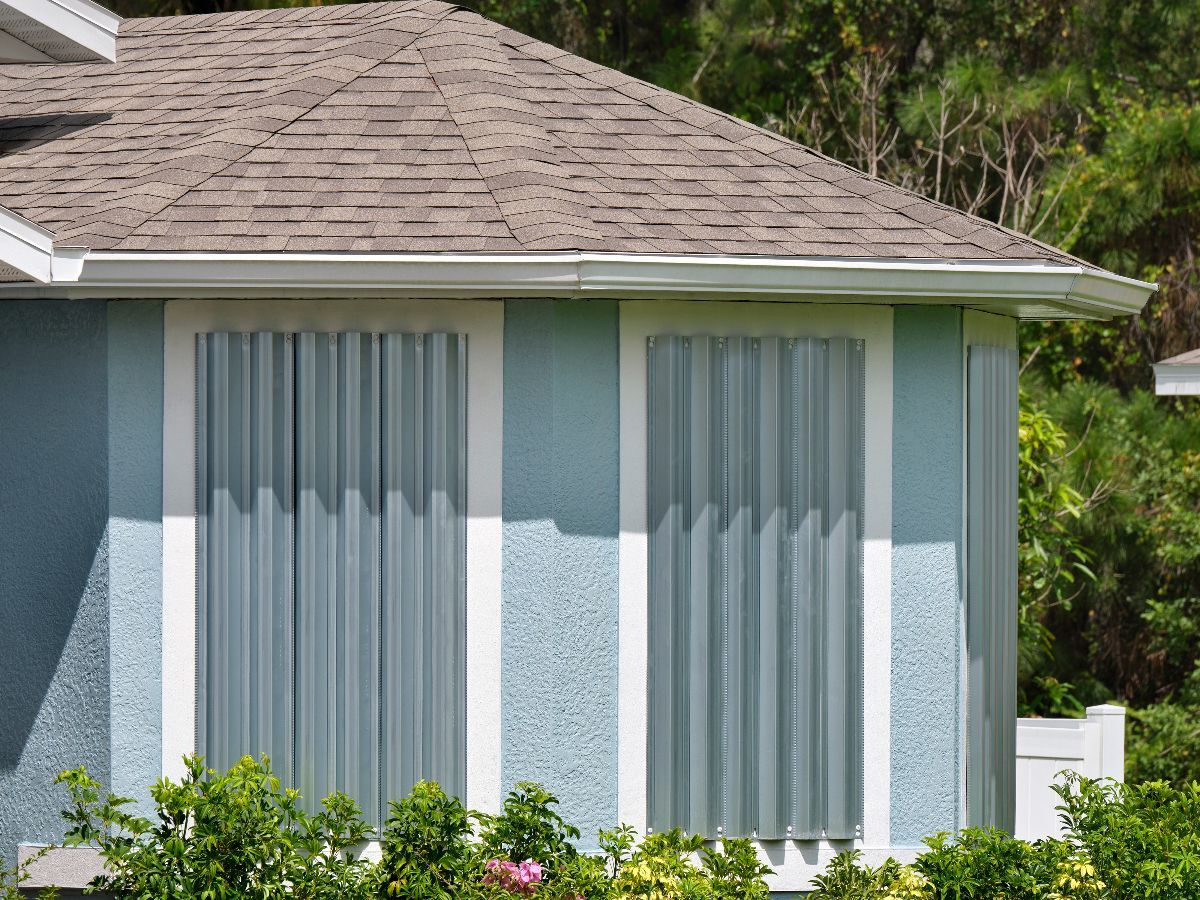 Blue house with hurricane shutters, brown roof, and green bushes.