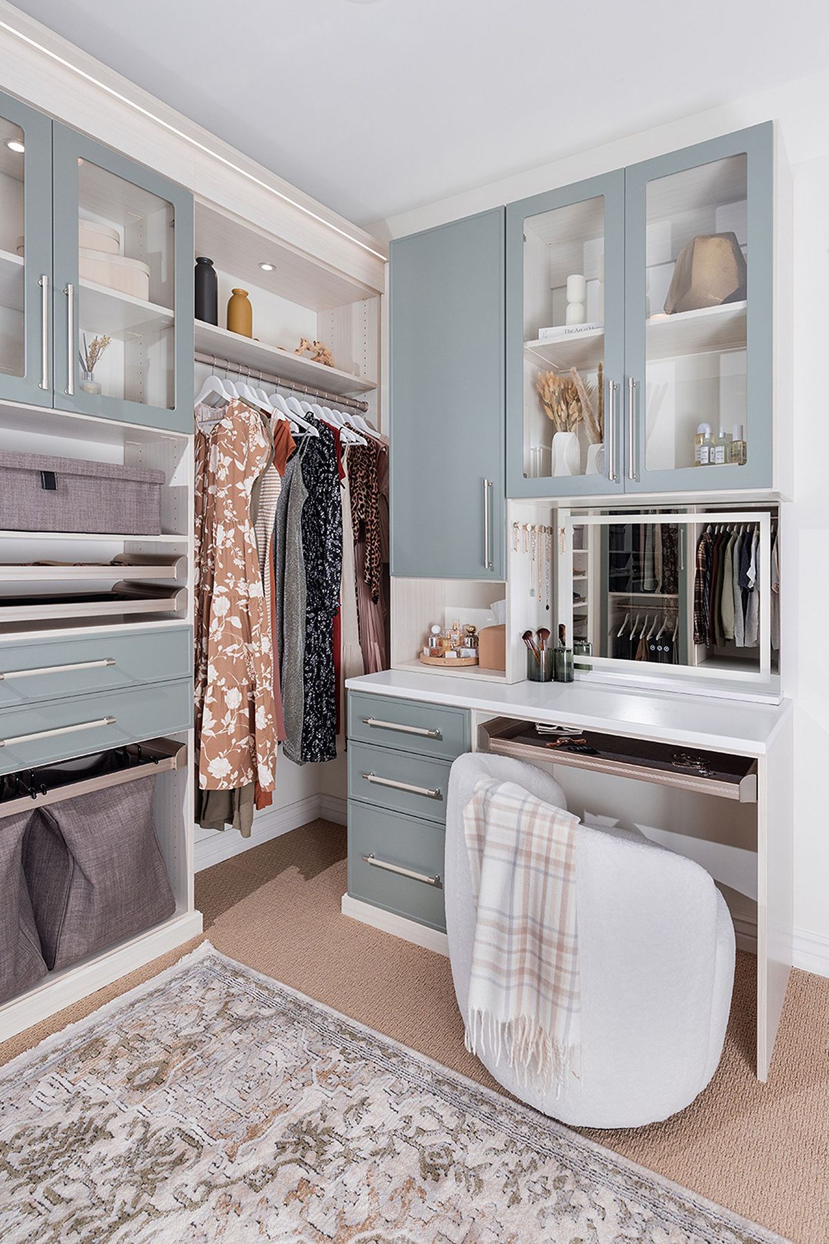 Walk-in closet with blue-gray cabinets, clothes, vanity with mirror, and white chair. Beige rug on floor.