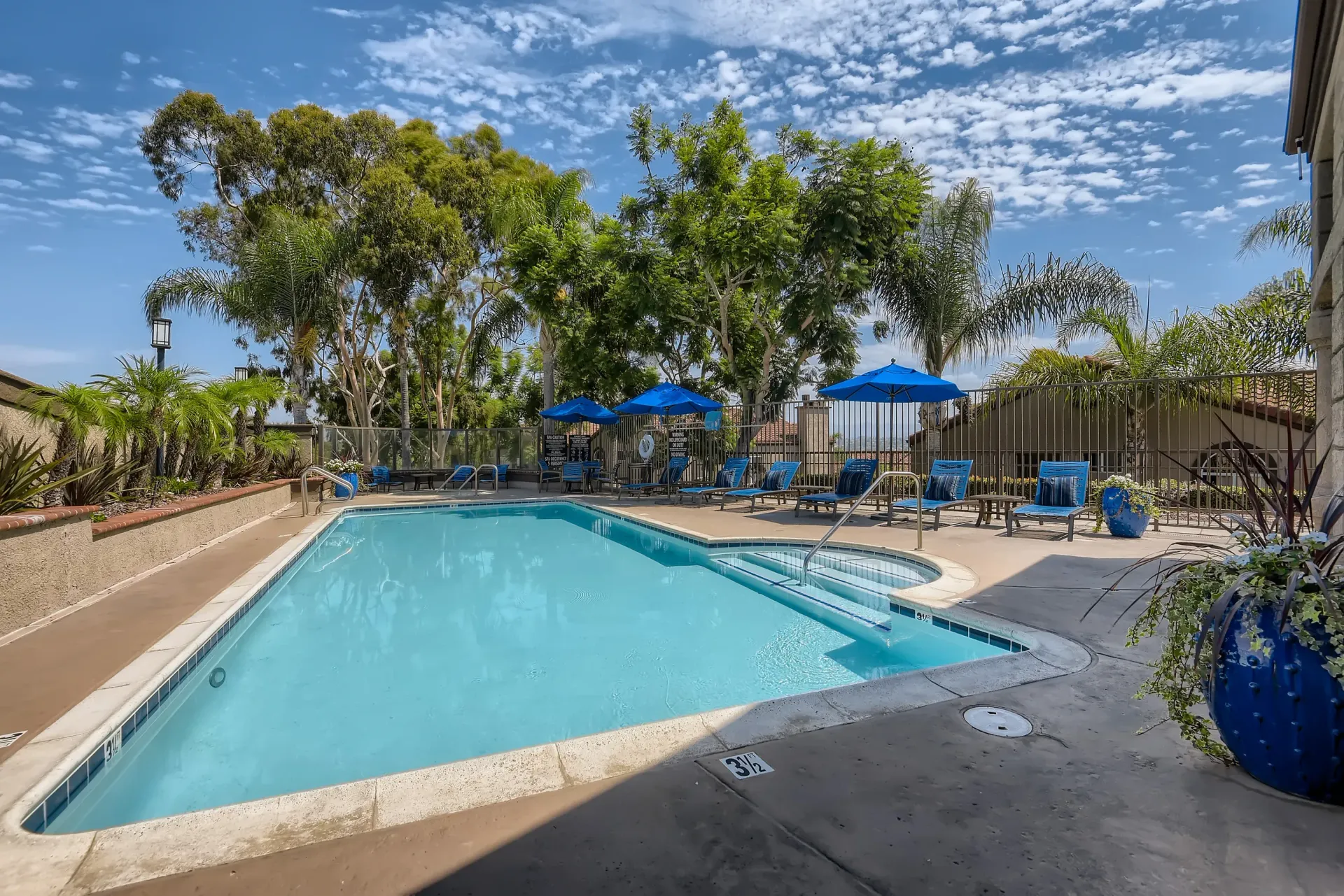 Outdoor community pool area with blue lounge chairs and umbrellas, surrounded by trees.