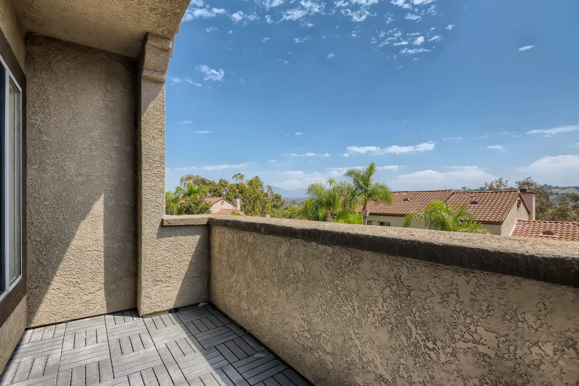 Balcony with beige stucco walls and railing, overlooking palm trees and red-tiled rooftops.