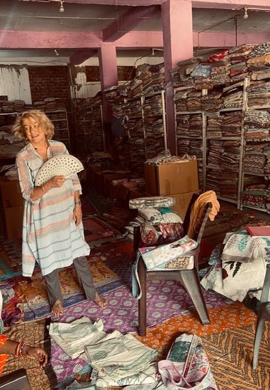 Donna in un magazzino di tappeti con in mano un articolo decorato, circondata da pile di tappeti e tessuti.