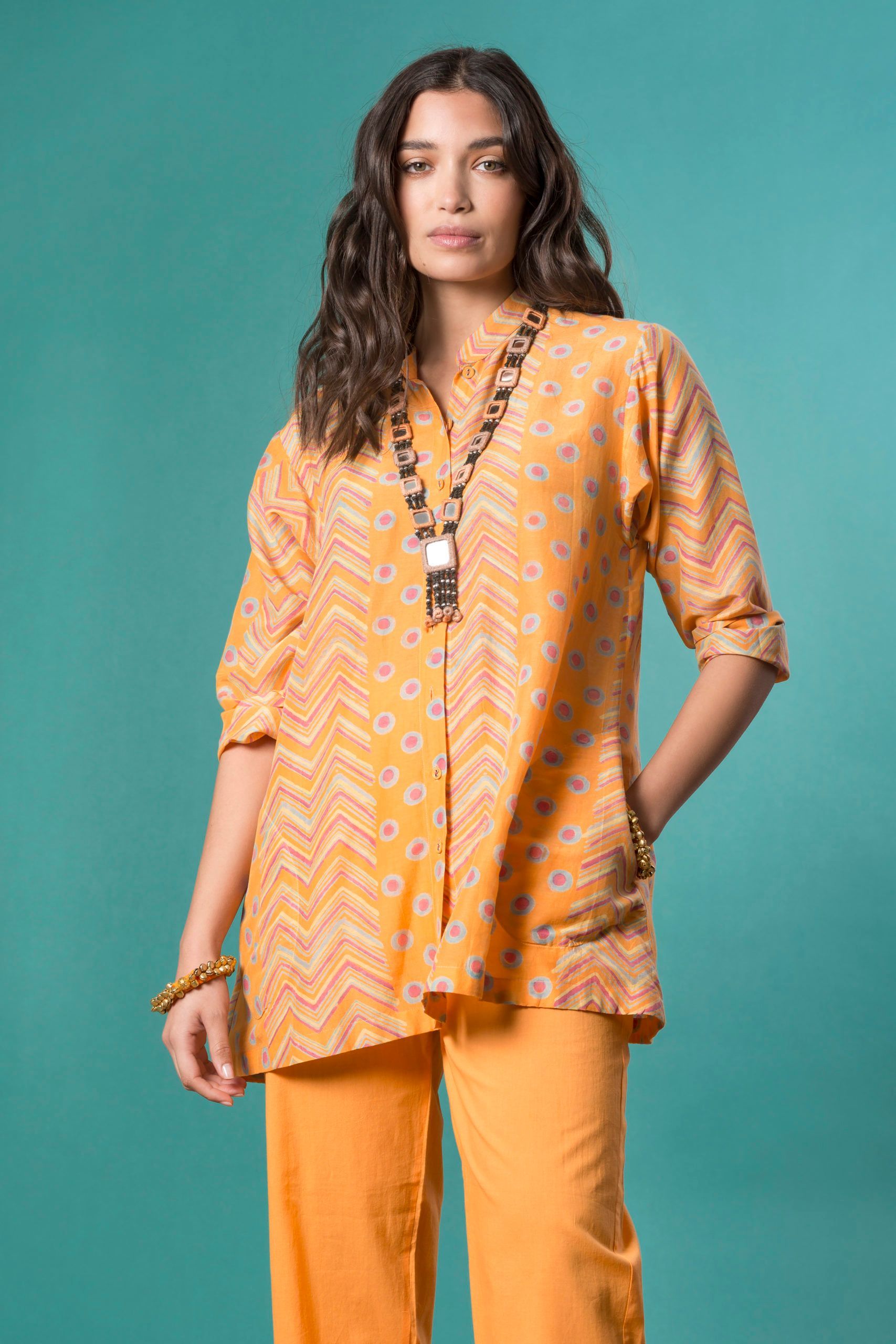 Donna con top e pantaloni a fantasia arancione, sfondo turchese, indossa una collana.