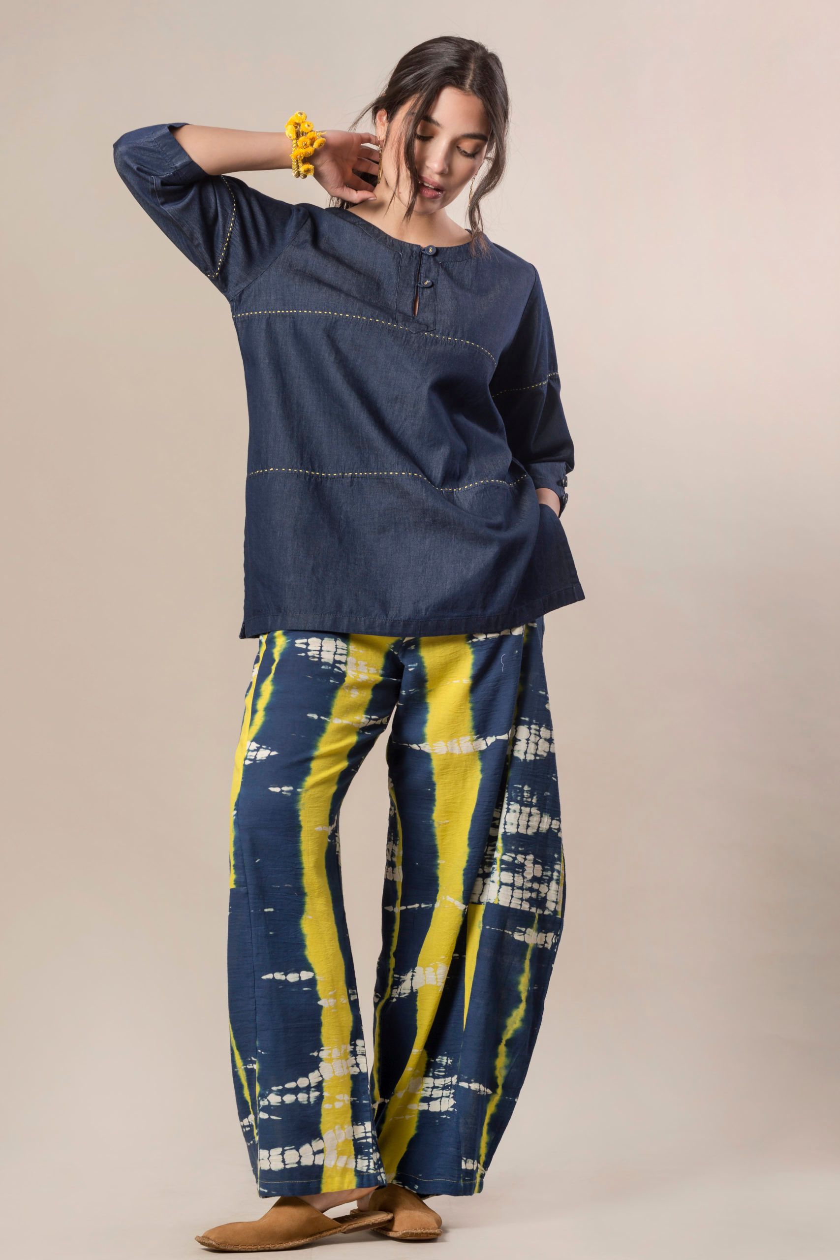 Donna con top in denim blu e pantaloni a fantasia blu e gialli, in posa con la mano vicino al viso.