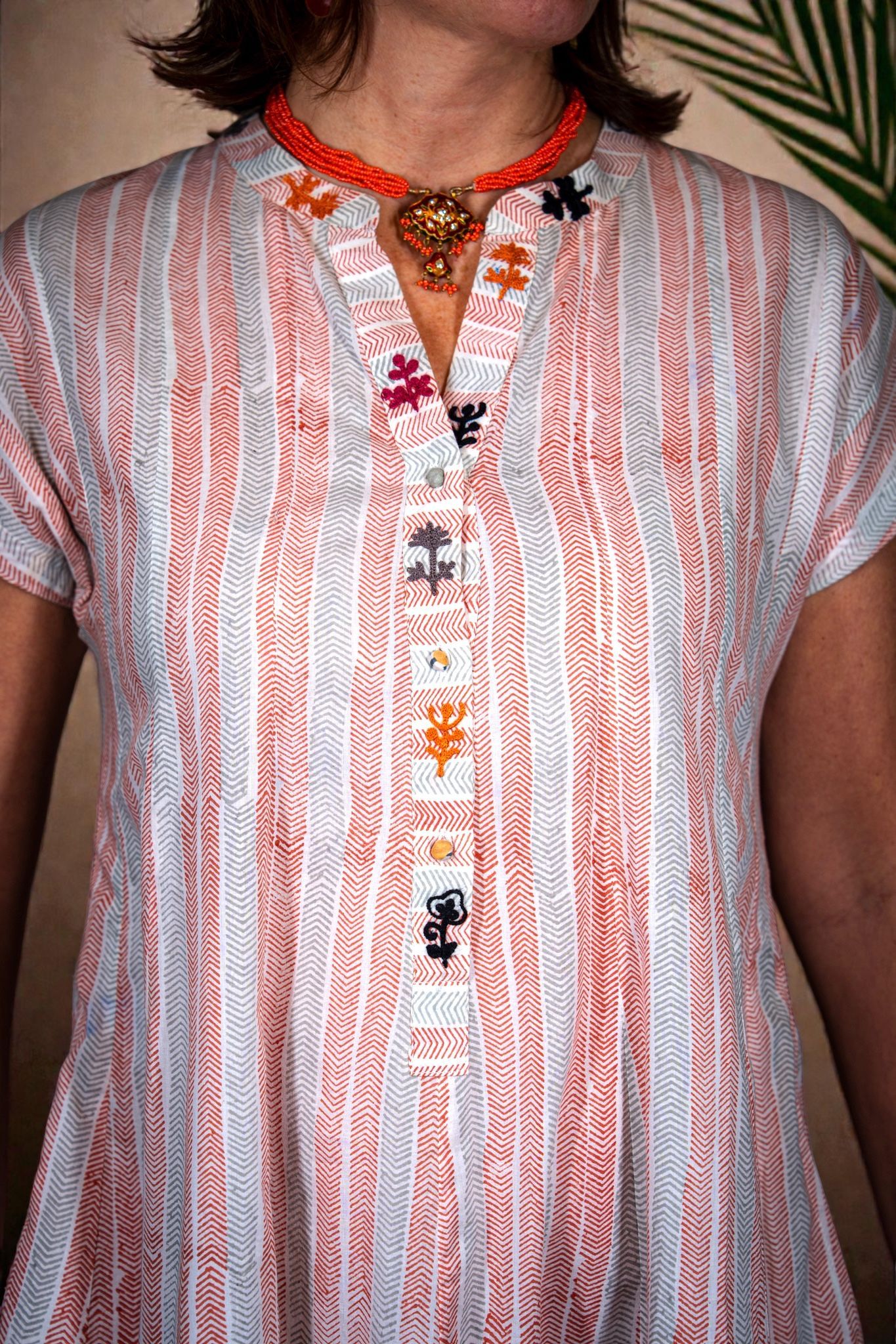 Una persona che indossa una tunica a maniche corte con una fantasia a righe verticali nei toni del corallo e del blu, e una collana di perline.
