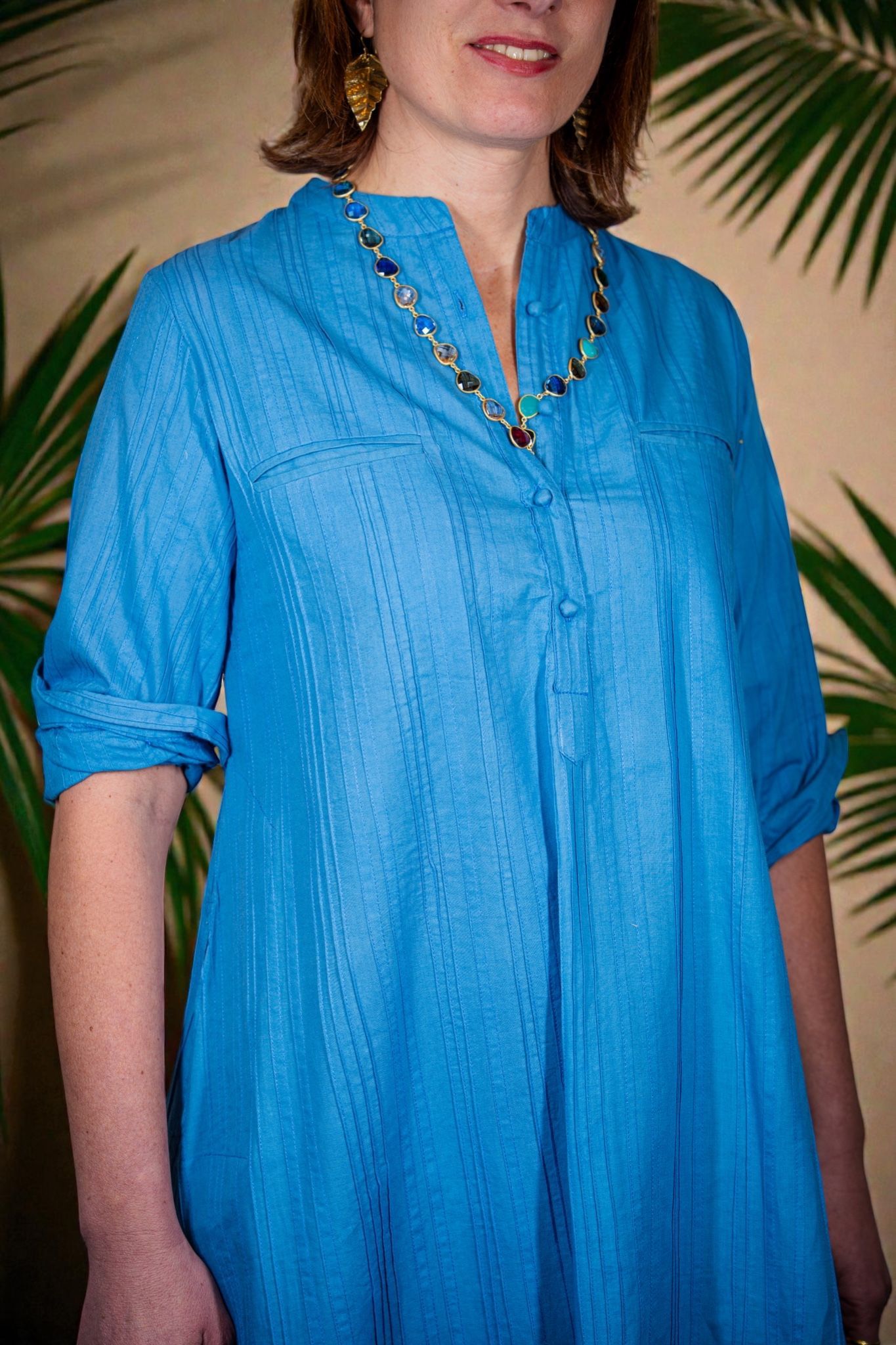 Abito blu brillante a maniche lunghe con scollo a V e una collana di perline, sullo sfondo di foglie di palma.