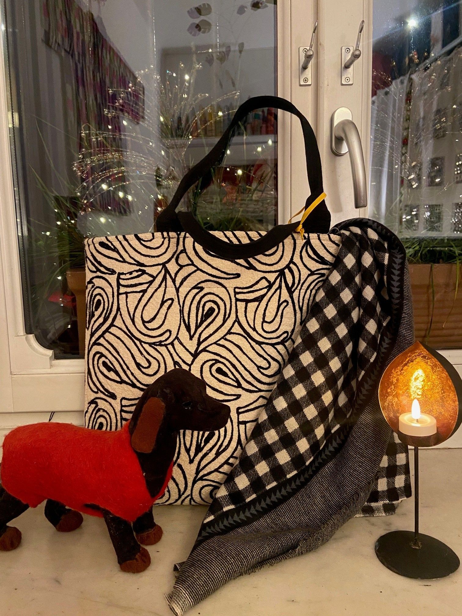 Borsa tote con motivo bianco e nero, una sciarpa a quadri, una statuetta di bassotto e una candela accesa sul davanzale di una finestra.