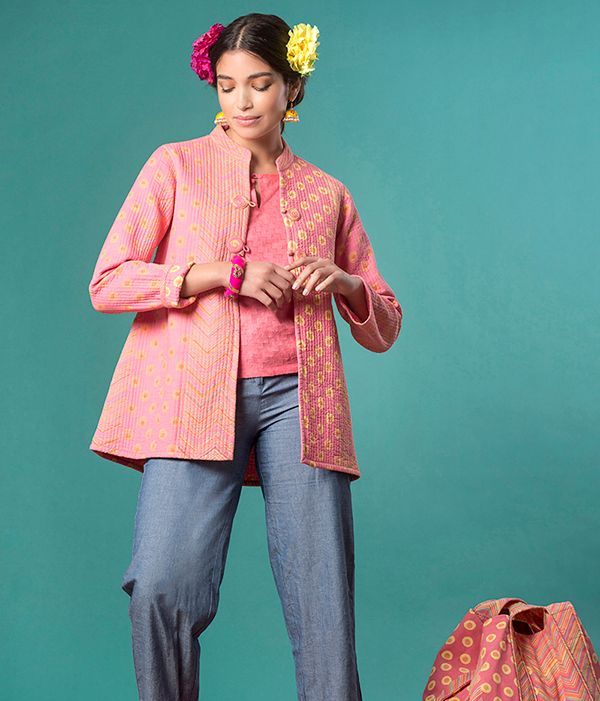 Donna con giacca rosa e pantaloni blu, in piedi su sfondo verde acqua, con in mano una borsa.