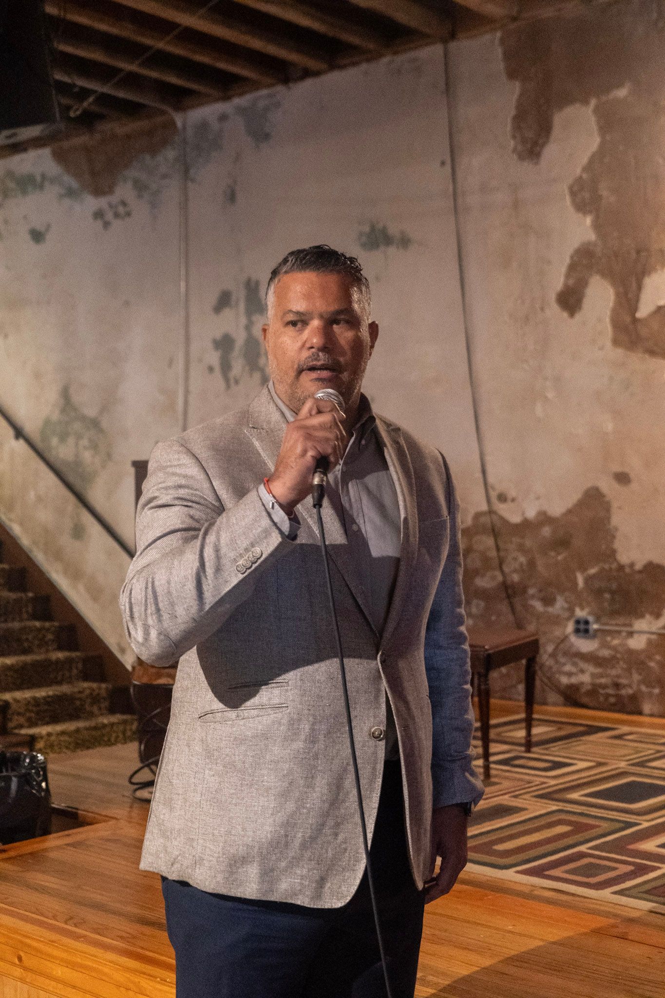 Man speaking into a microphone, wearing a light-colored blazer, standing indoors near stairs and distressed walls.