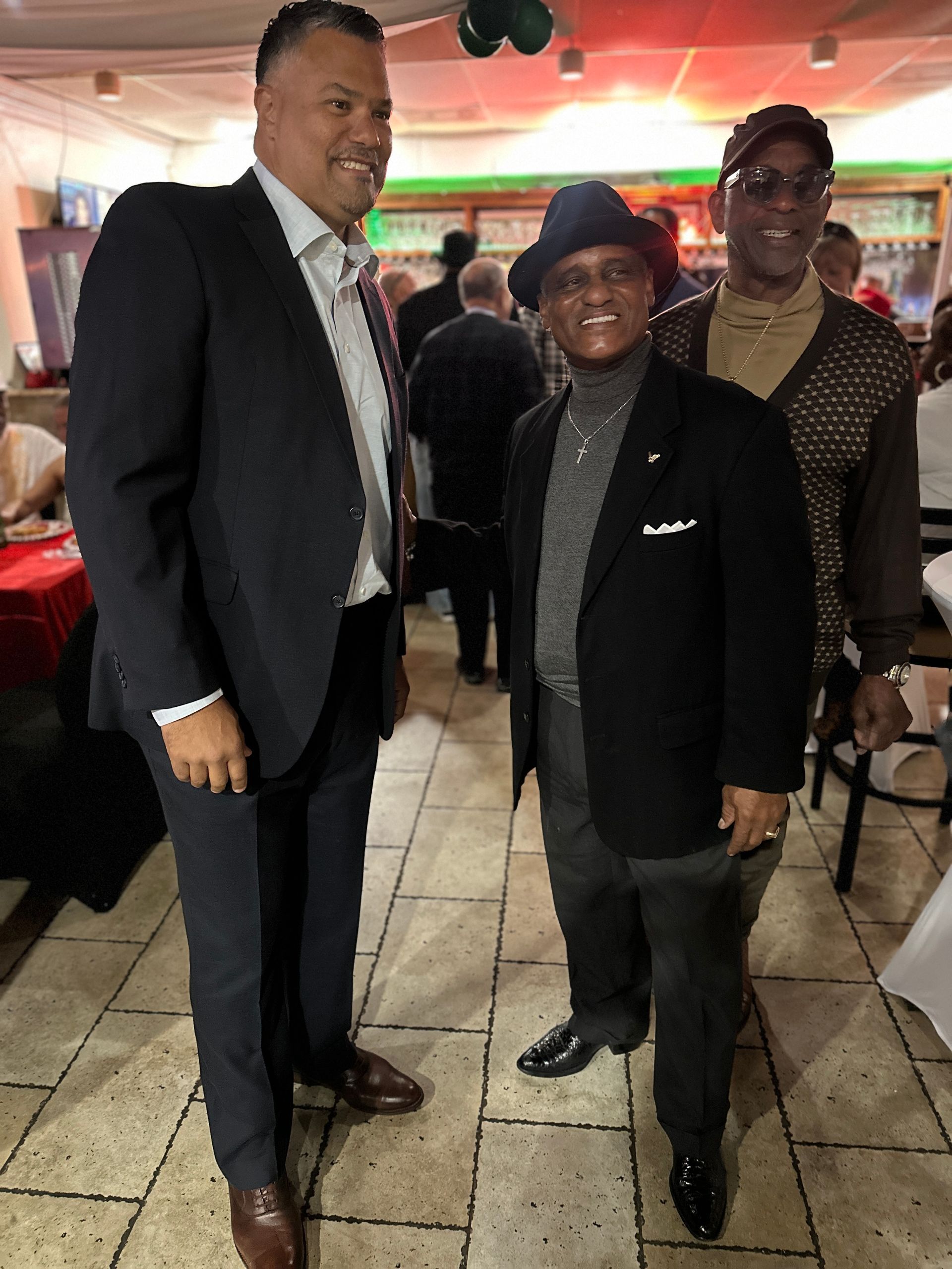 Three men standing inside, possibly at an event. The men are wearing suits, and one has a hat.