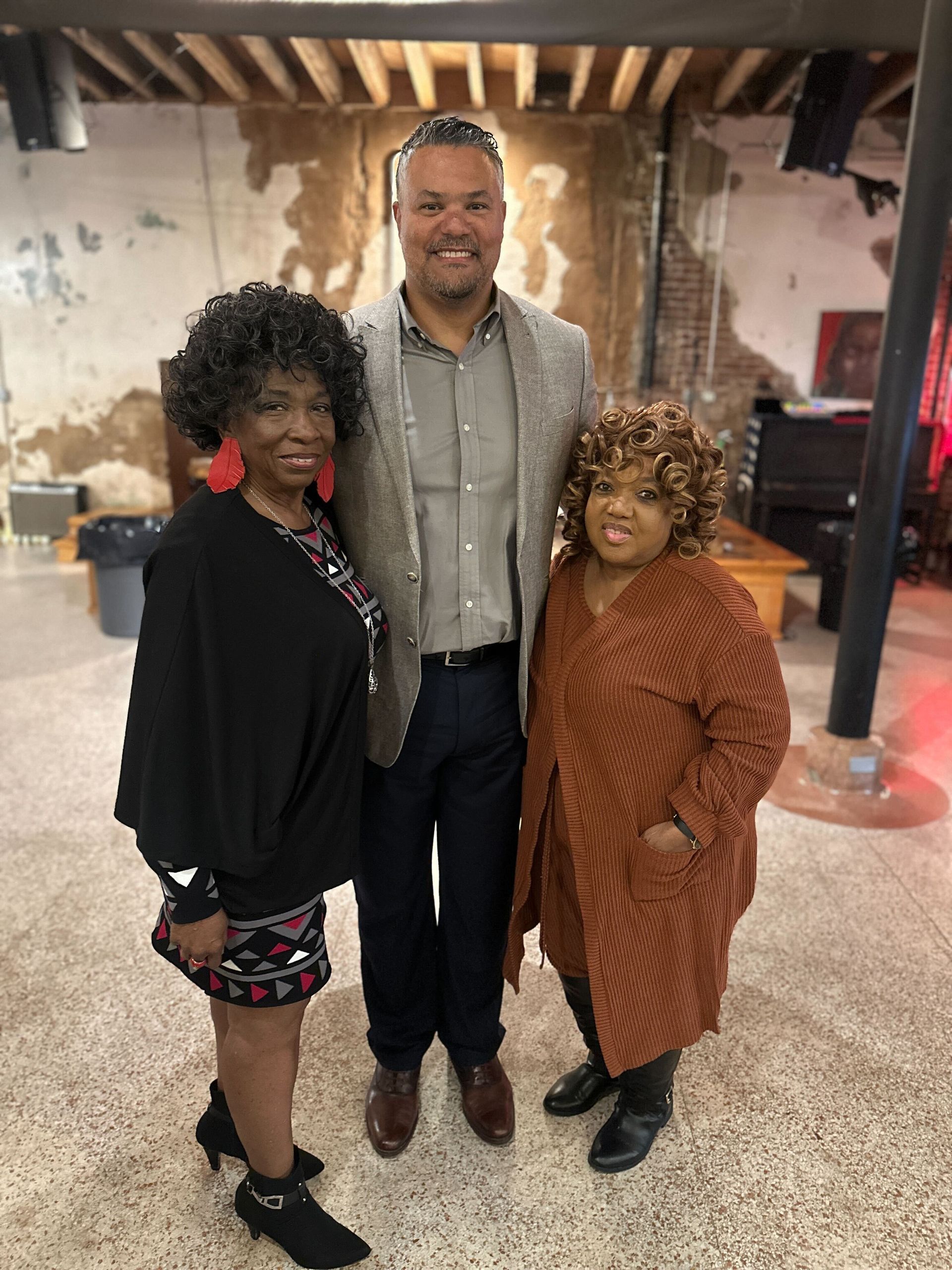 Woman and man smiling, posing indoors. Man in gray blazer. Woman in black top. Dimly lit setting.