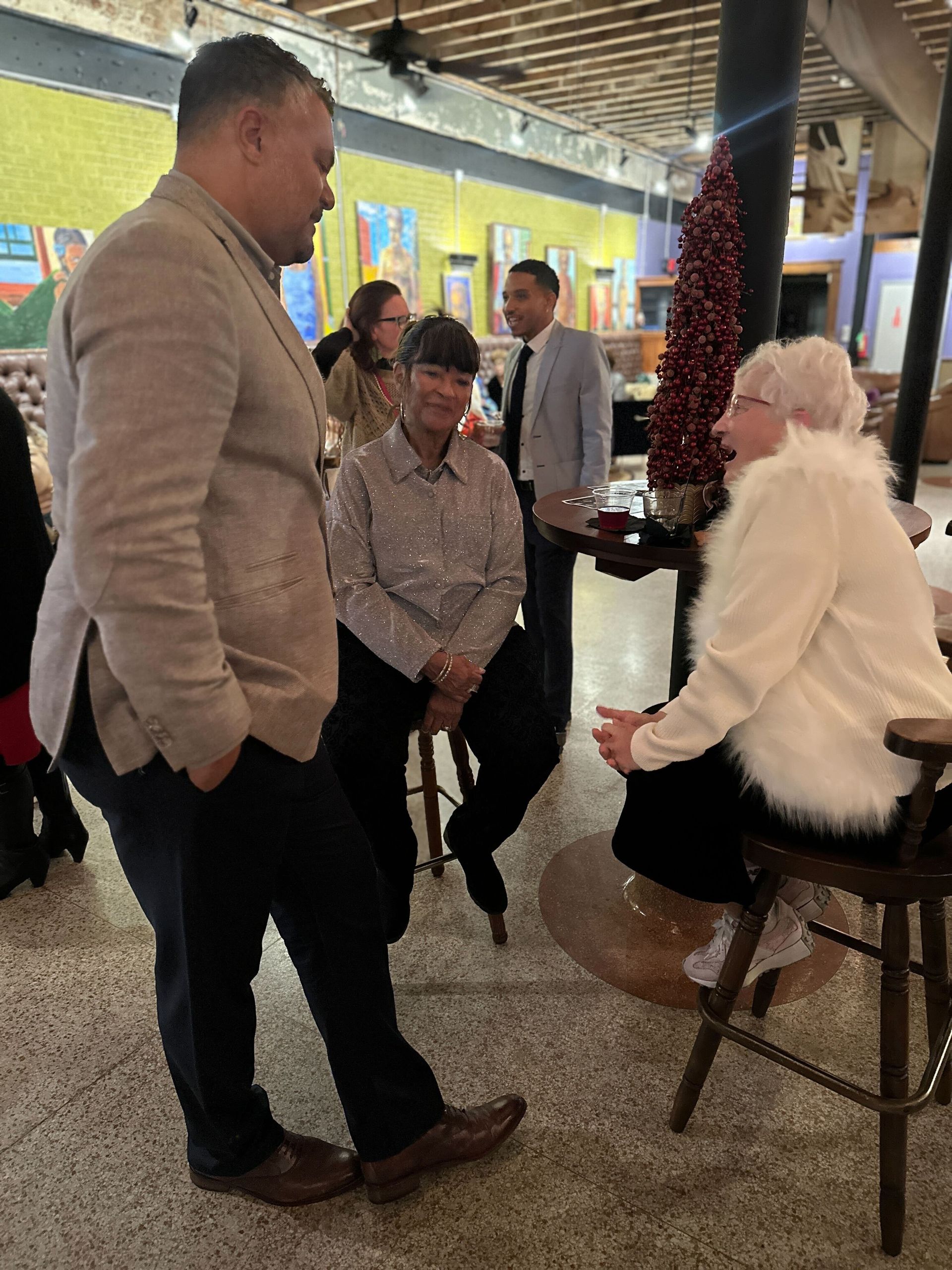 People at a gathering inside; a man in a blazer talks to an elderly person seated, another person sits nearby.