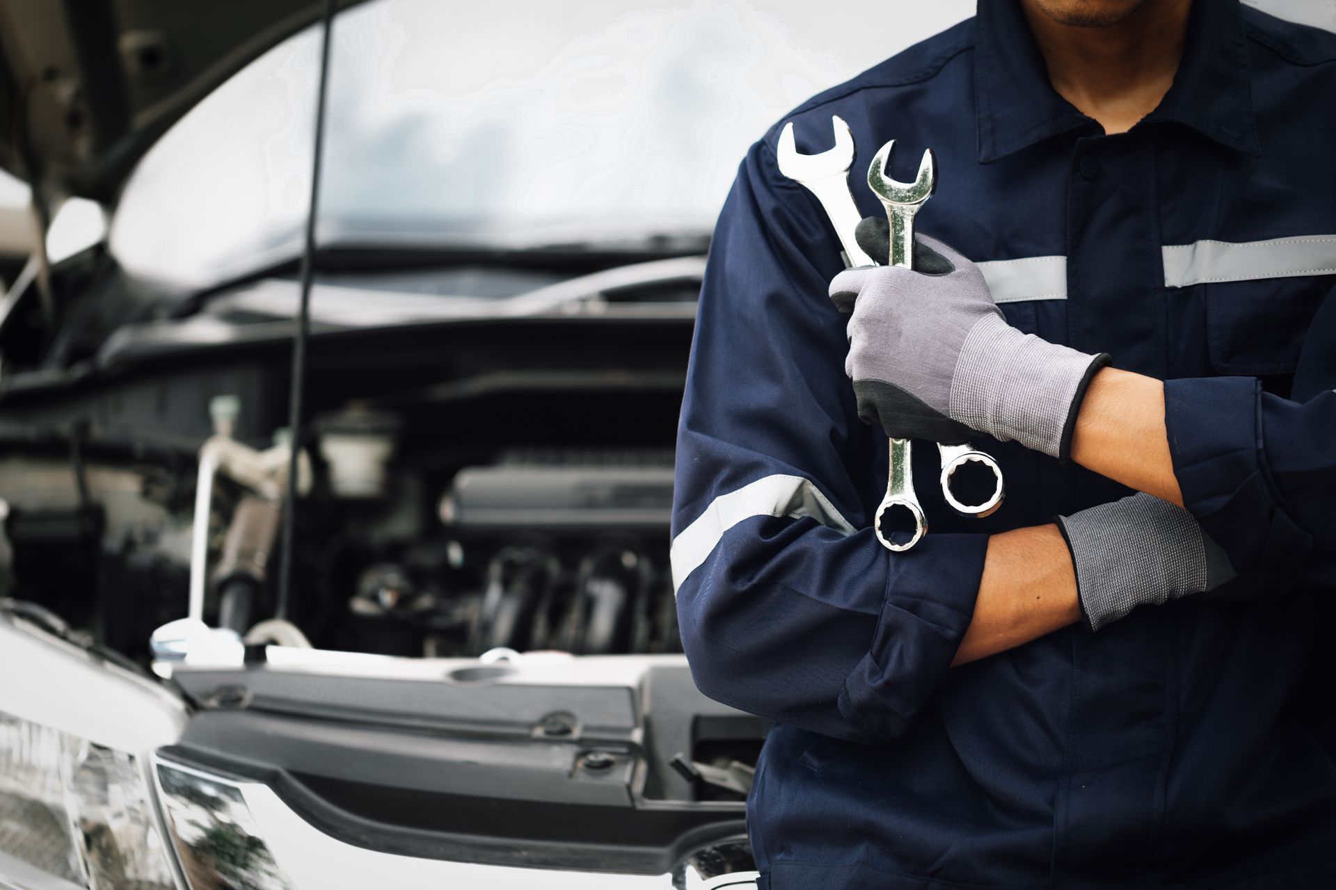 Meccanico con chiavi inglesi davanti al cofano aperto di un'auto. Uniforme da lavoro blu, guanti grigi.