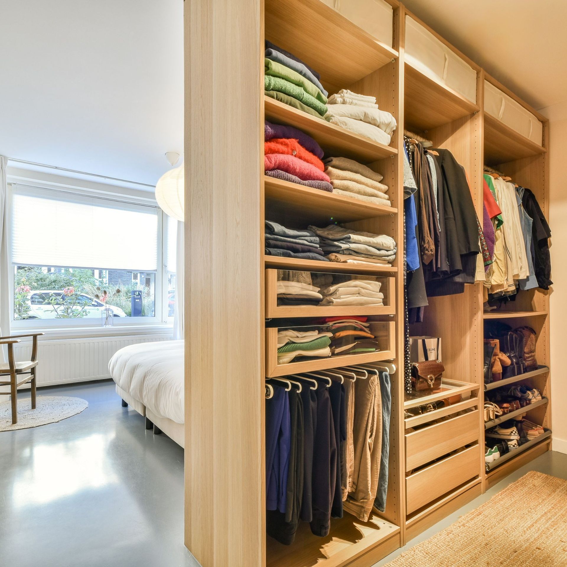 Wooden closet unit with clothes organized, next to a bed in a room with a window.