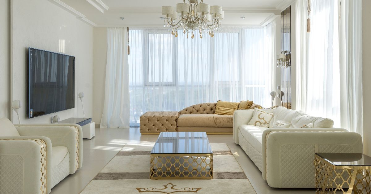 Elegant living room with white and gold furniture, a large TV, and sheer white curtains.