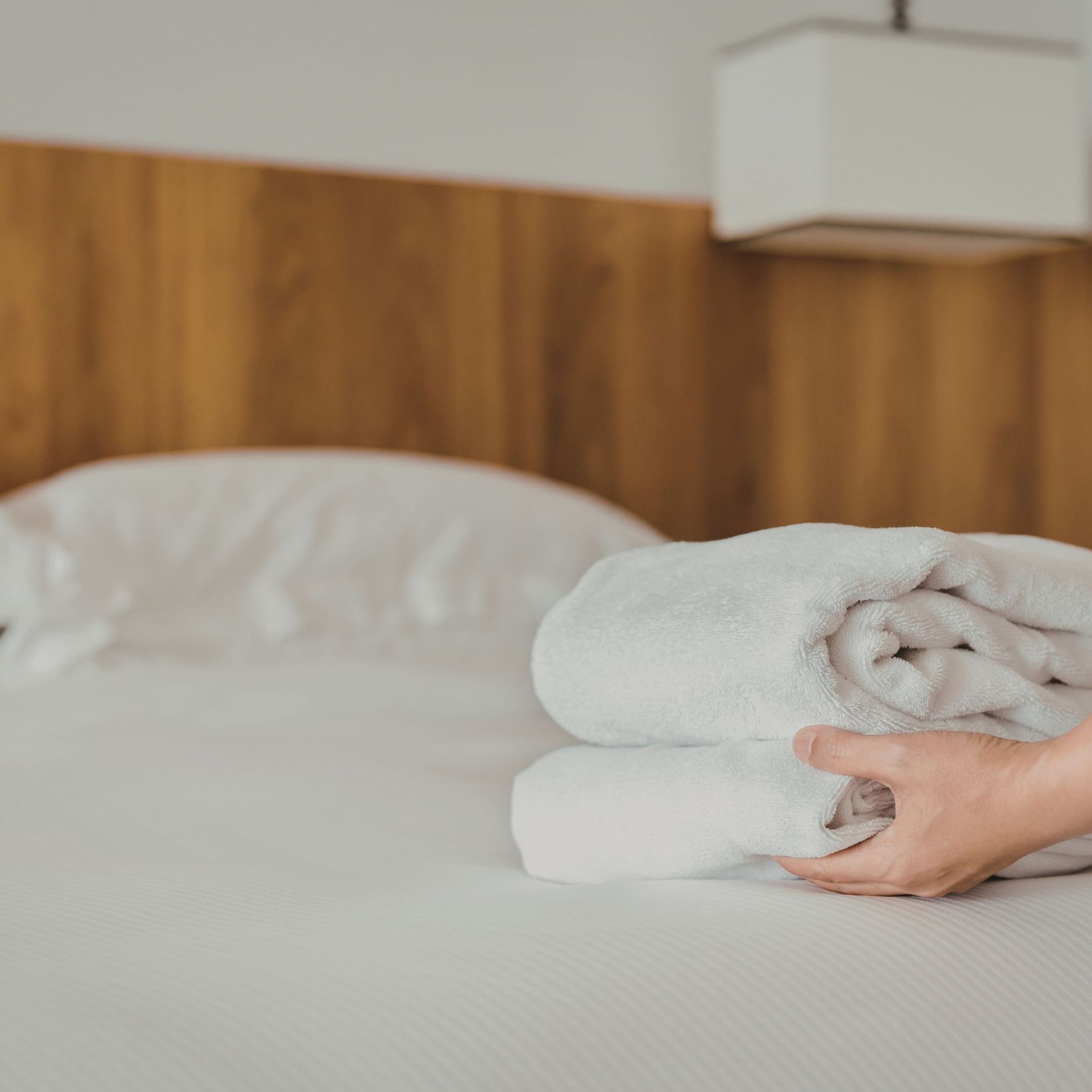 A hand placing folded white towels on a bed with a white pillow; wooden headboard in background.