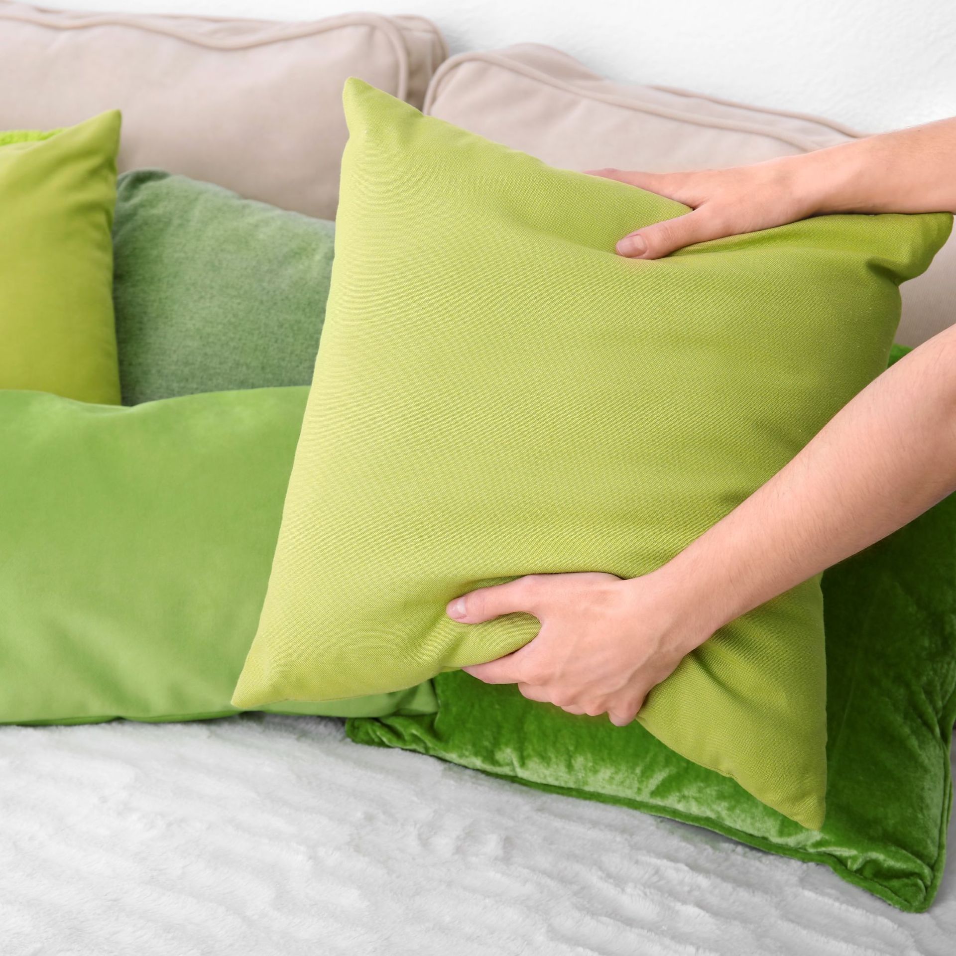 Hands arranging a lime-green pillow on a sofa with several green pillows of different shades.