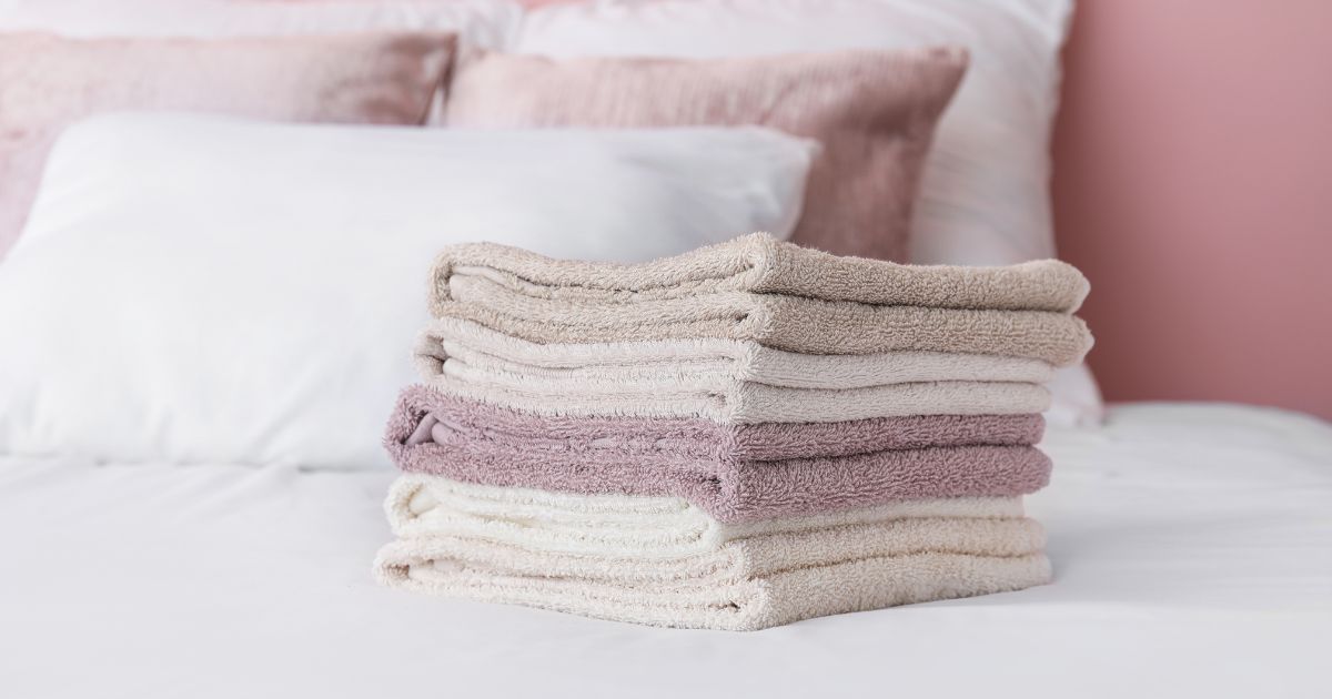 Stack of folded beige and mauve fluffy towels on a white bed with pink pillows.