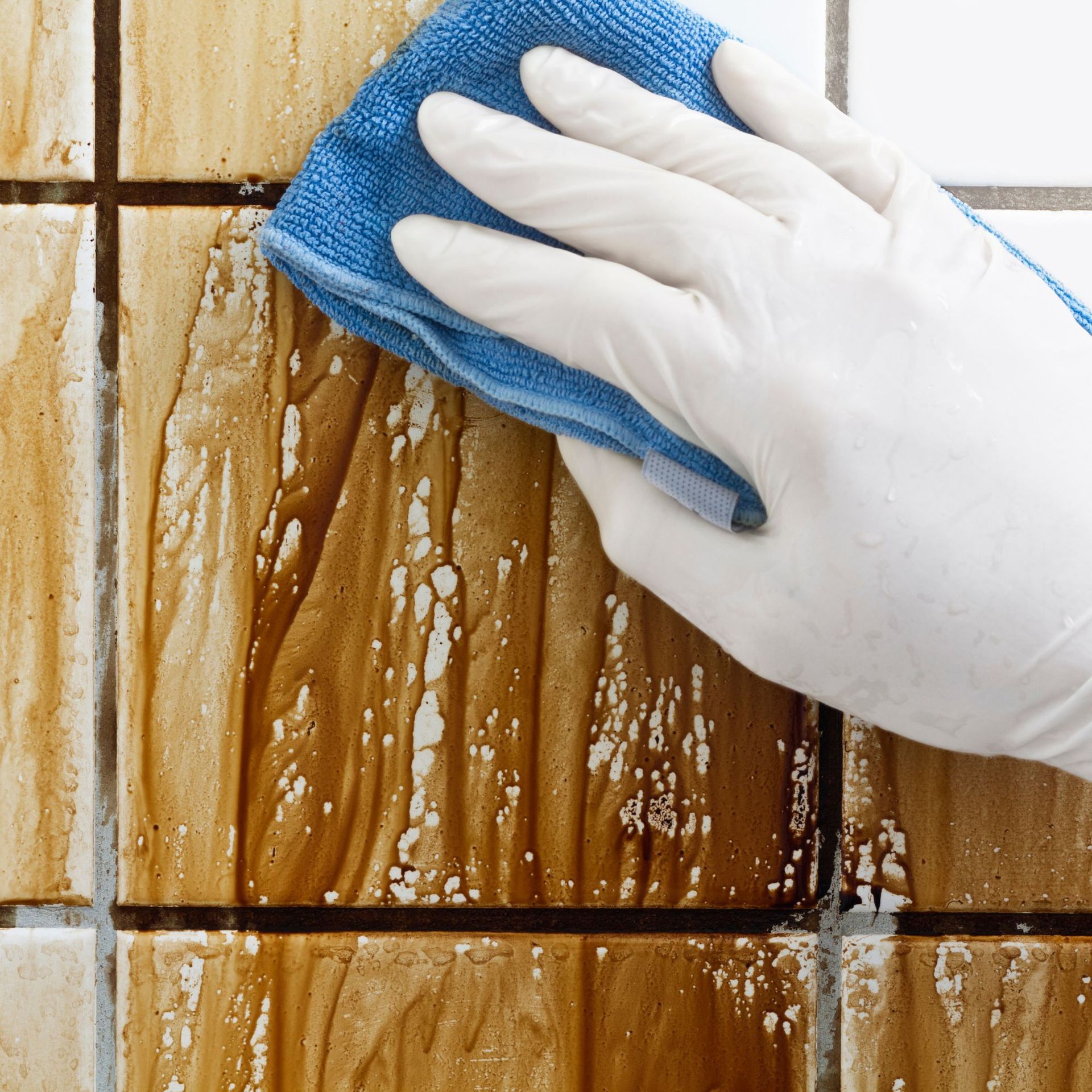 Hand in a white glove wiping a brown-stained tile wall with a blue cloth.