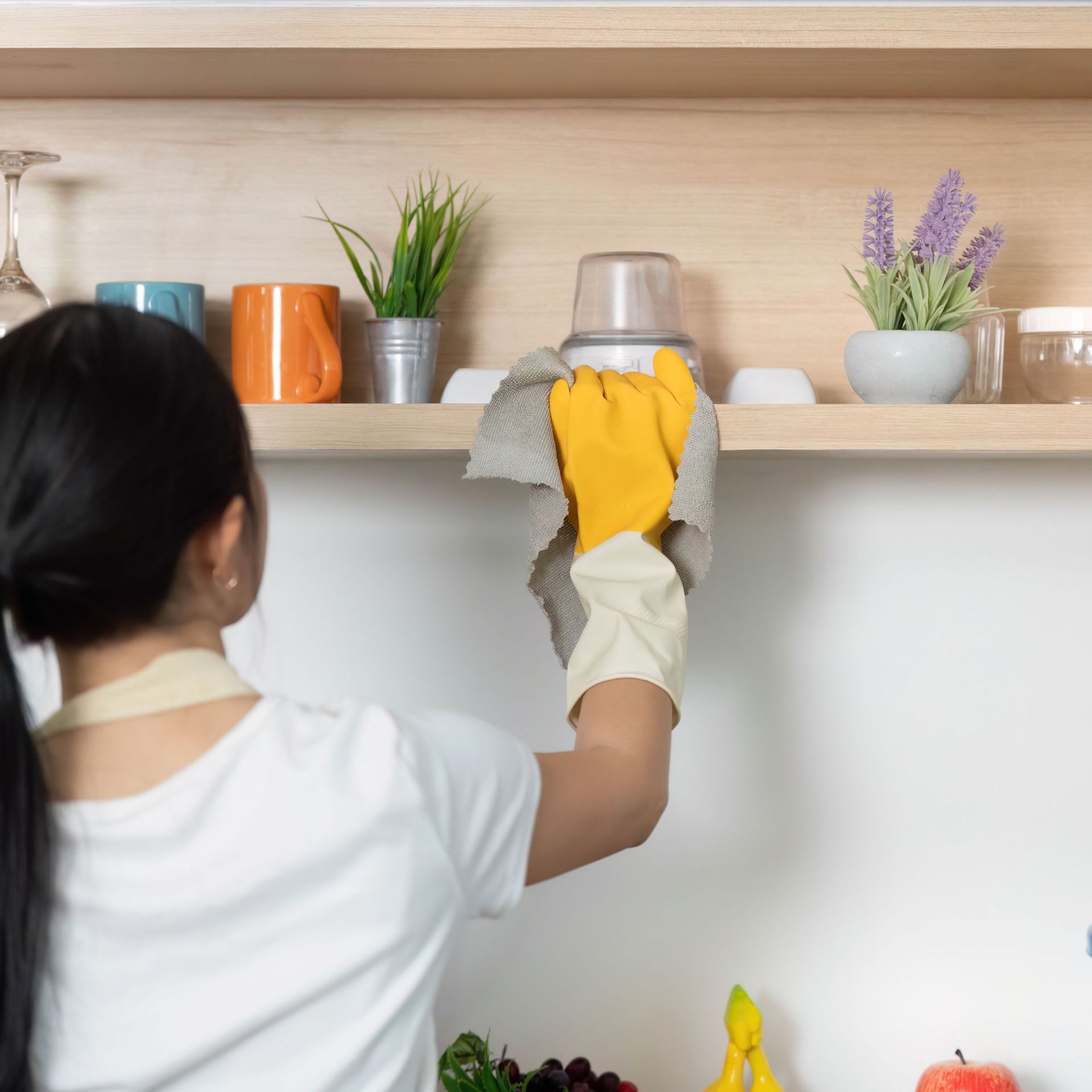 Woman cleaning a kitchen shelf with a yellow-gloved hand, other hand in white glove. Shelf has mugs, plants.