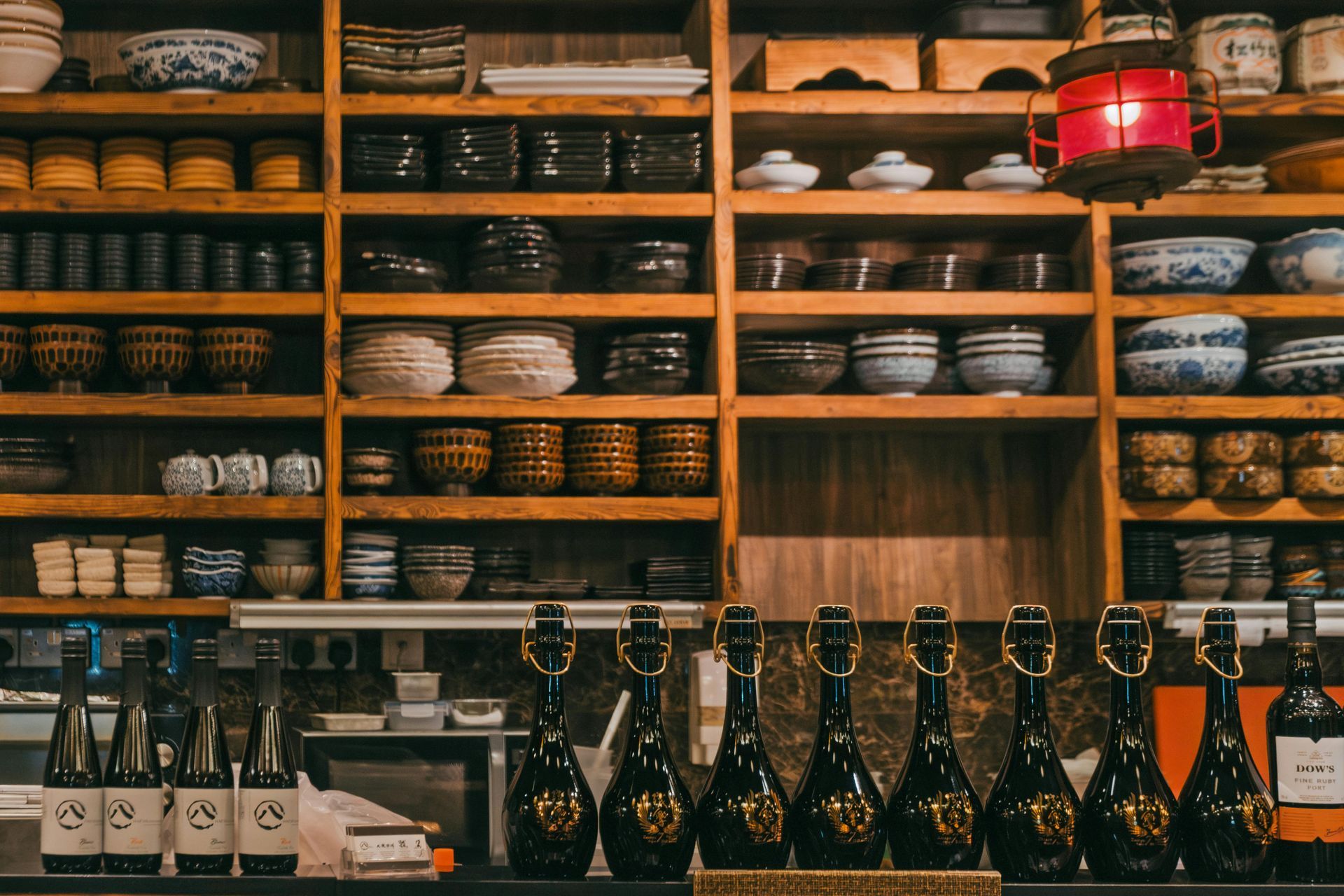 Shelves of pottery behind a bar, displaying bottles, with a red lantern hanging above.