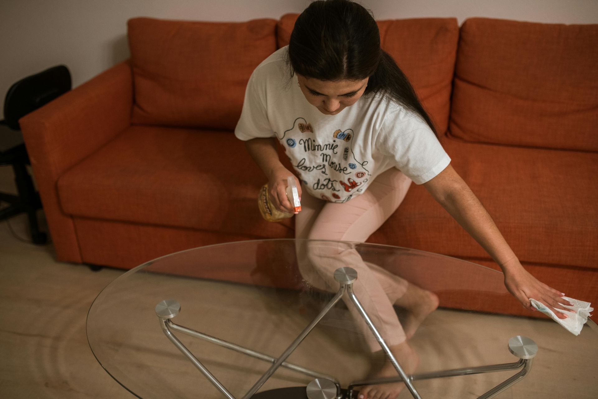 Woman cleaning a glass coffee table with spray bottle and cloth, near an orange sofa.