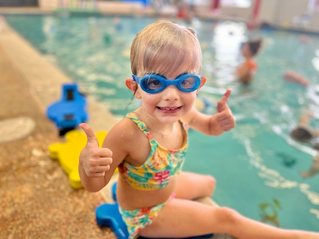 A little girl wearing goggles is giving a thumbs up in front of a swimming pool.
