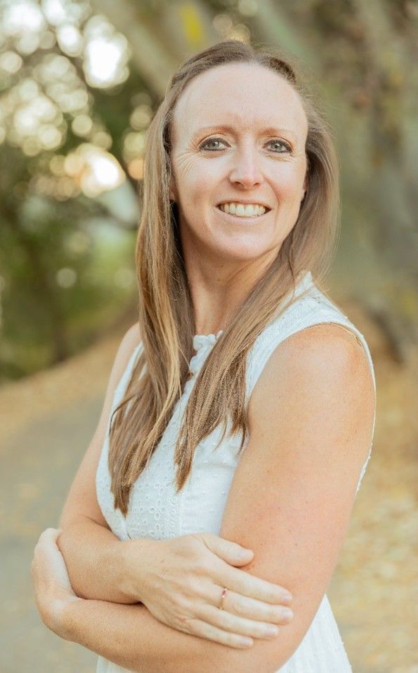 A smiling person with long light brown hair wearing a white sleeveless top, standing outdoors with arms crossed.