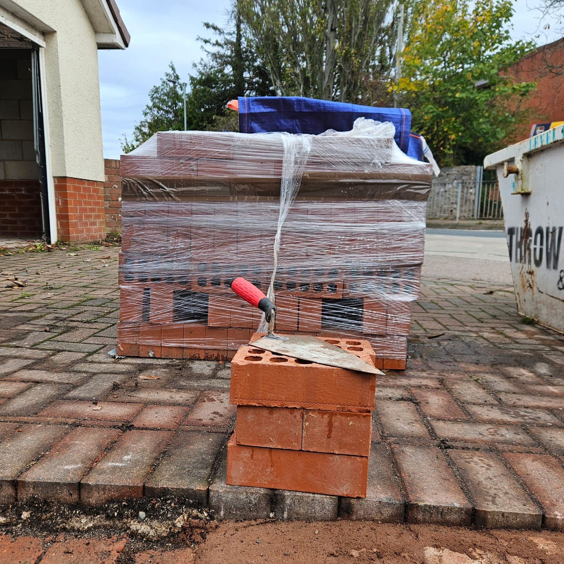 Bricks stacked on a brick walkway with a trowel, a pallet of wrapped bricks, and a dumpster.