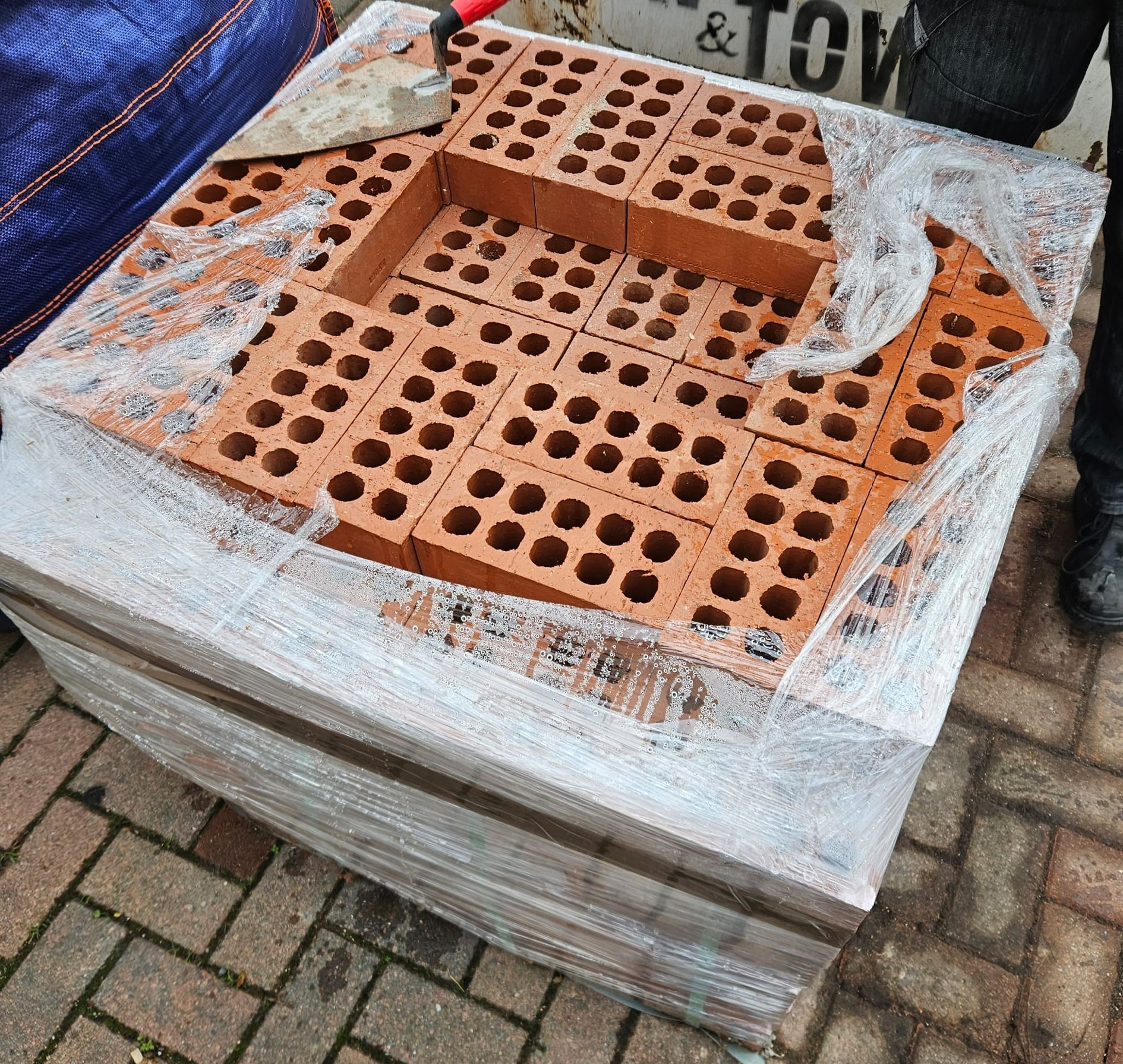 Pallet of red bricks wrapped in clear plastic, outdoors on brick pavers.